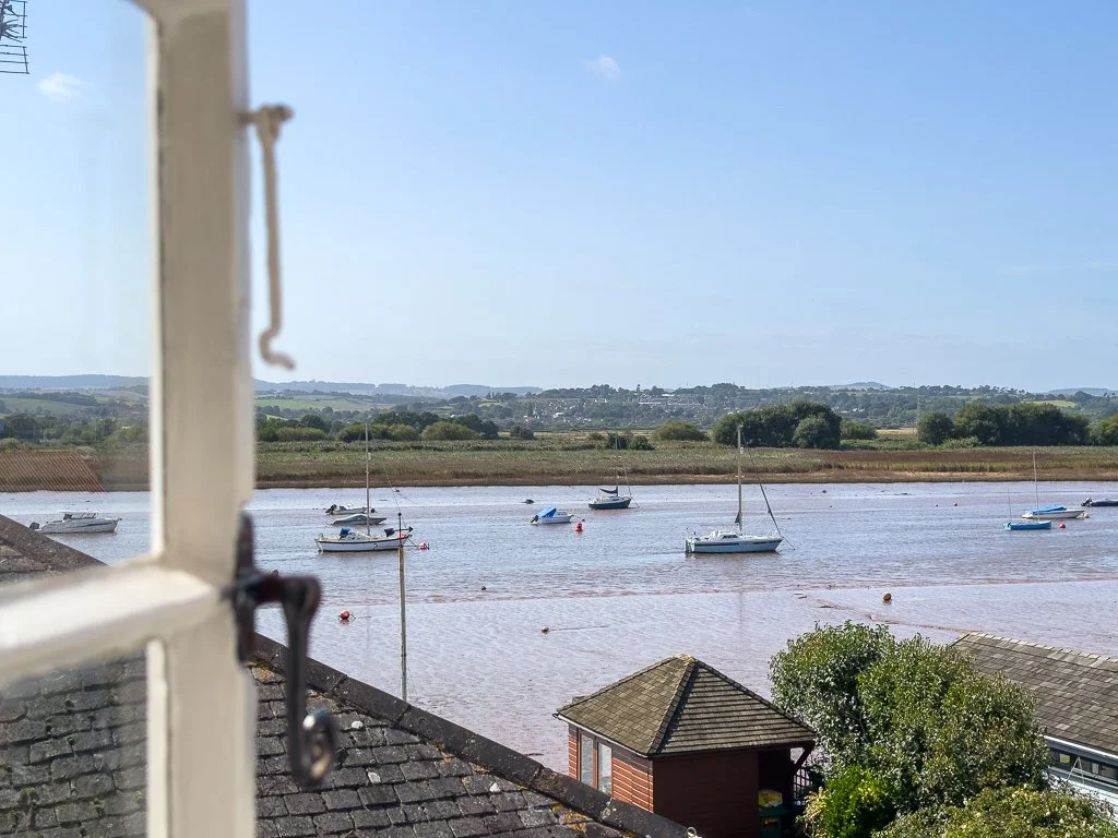 View of a river with several sailboats anchored, seen through a white-framed window. There are rooftops and greenery in the foreground with a distant landscape under a clear blue sky.