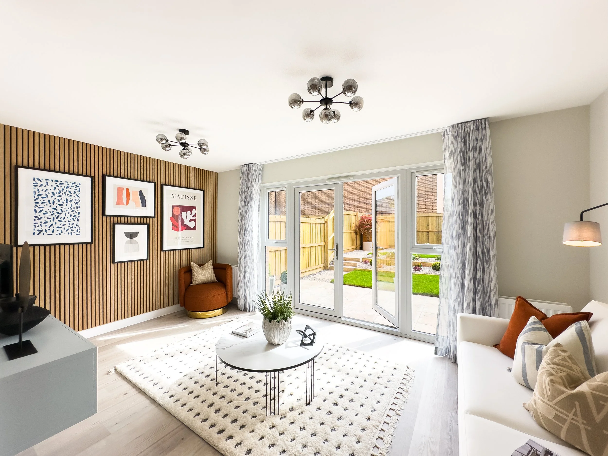 Bright living room with white walls, wooden accent wall with framed artwork, large sliding glass doors with patterned curtains, white furniture, and modern ceiling lights.