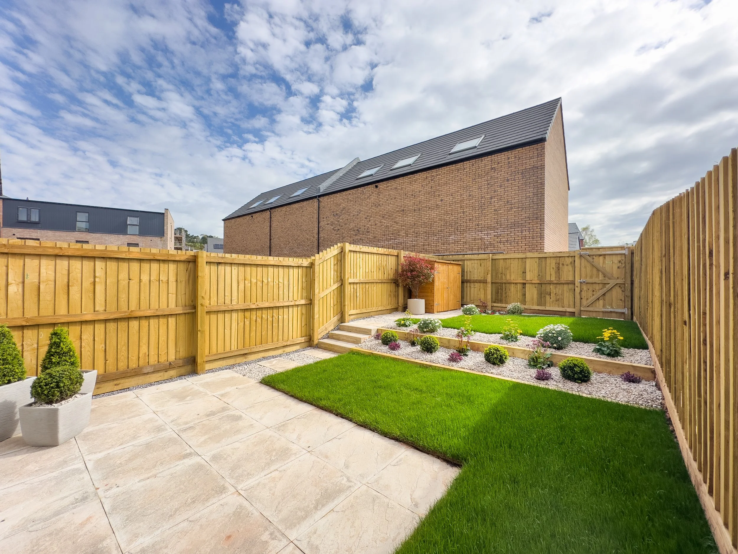 A backyard with a wooden fence, small potted plants, a green lawn, flower beds, and a brick house with skylights and a sloped roof under a partly cloudy sky.