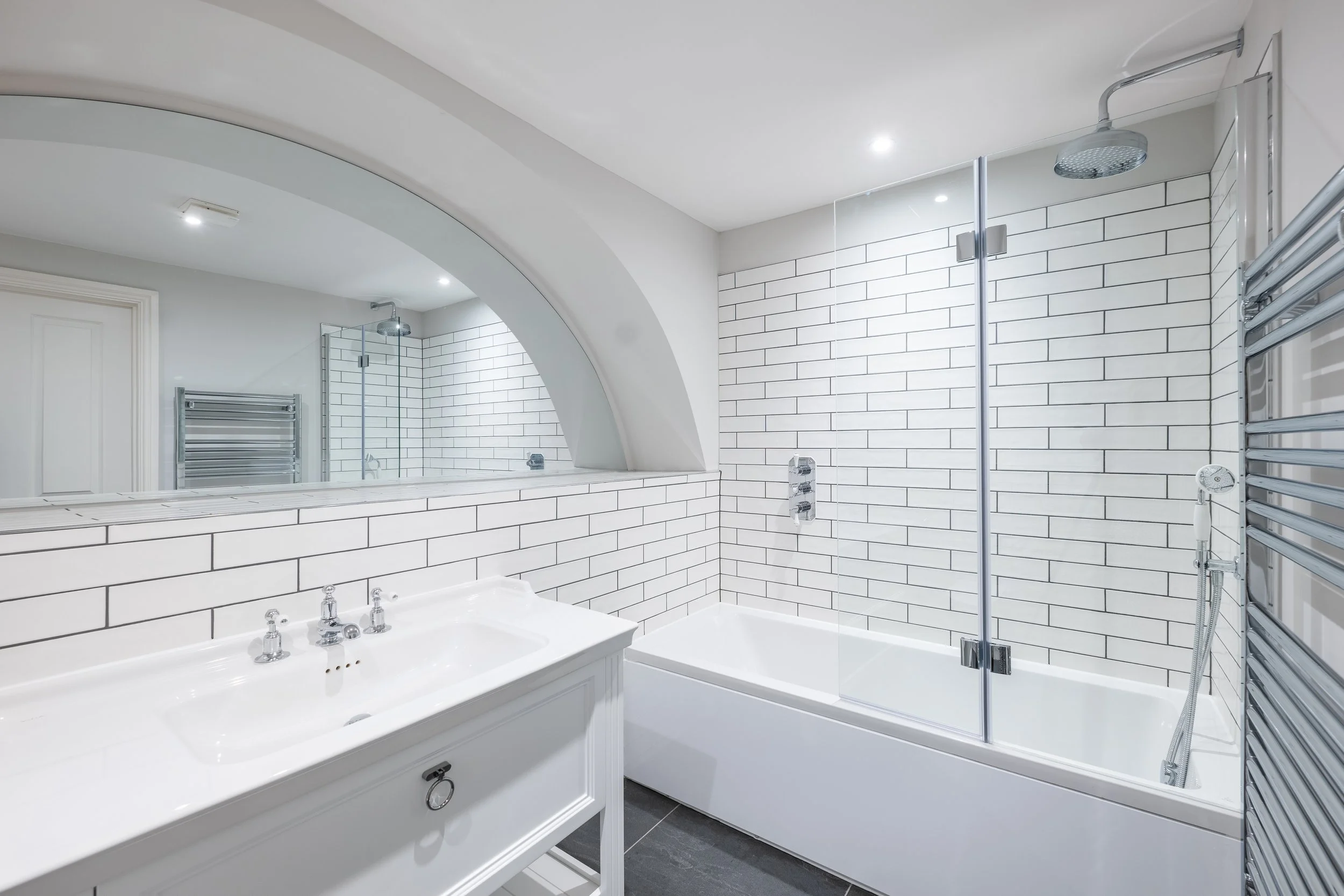 A modern bathroom with a white bathtub, white tiled walls, a walk-in shower with a rain shower head, a heated towel rack, and a vanity with a white sink.