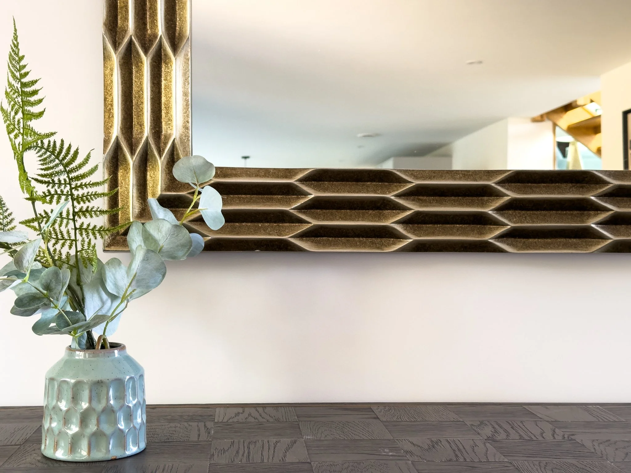 A tall ceramic vase with a ribbed texture and light green glaze, holding green fern and eucalyptus leaves, placed on a wooden floor with a mirror on the wall behind.