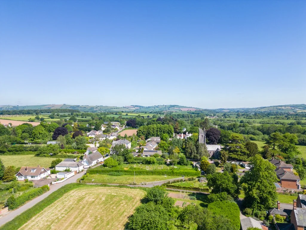 Aerial view of a small village surrounded by green fields and trees under a clear blue sky.