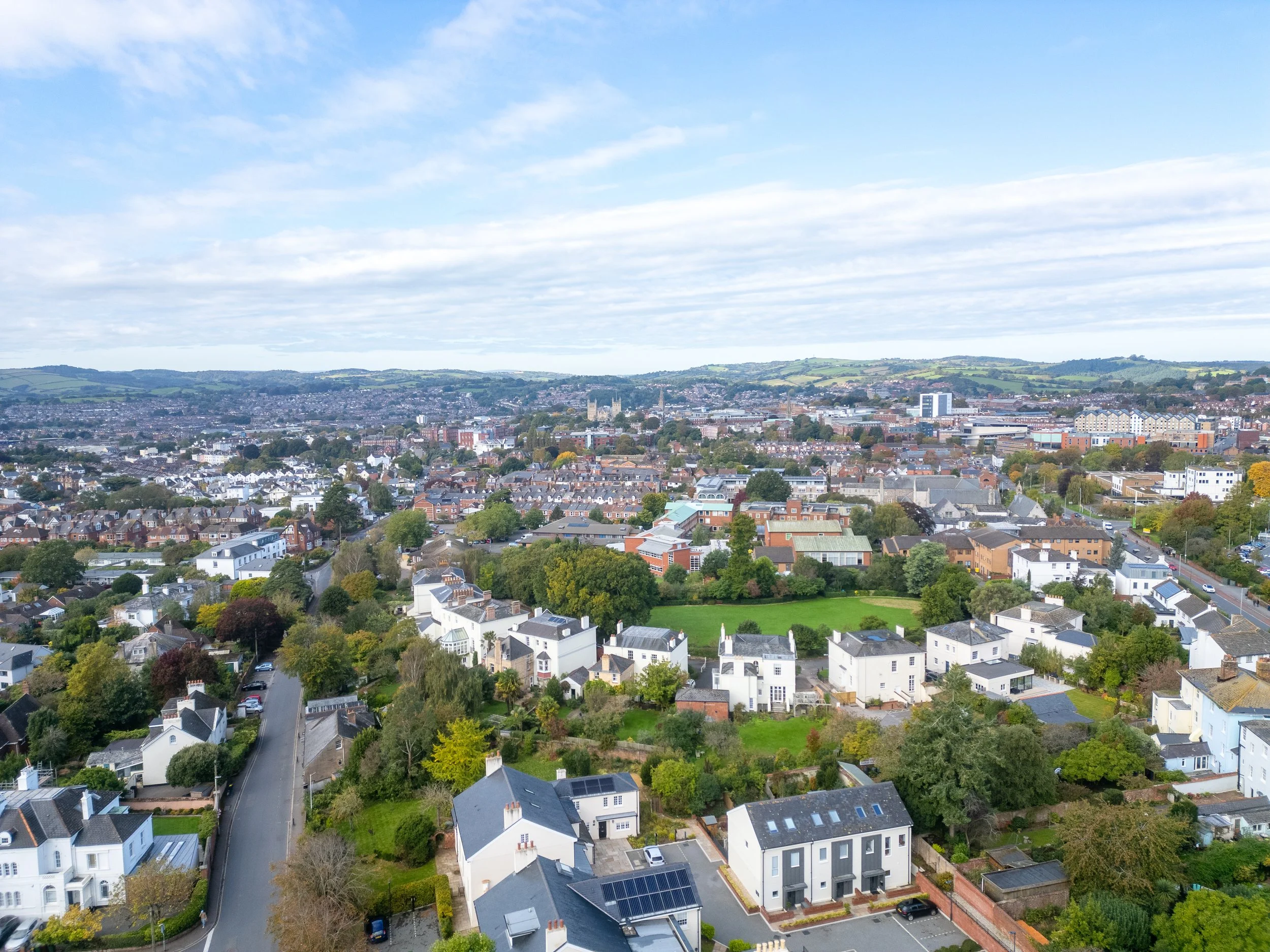 Aerial view of a suburban cityscape with residential houses, streets, and a few commercial buildings, with green hills in the background and partly cloudy sky.