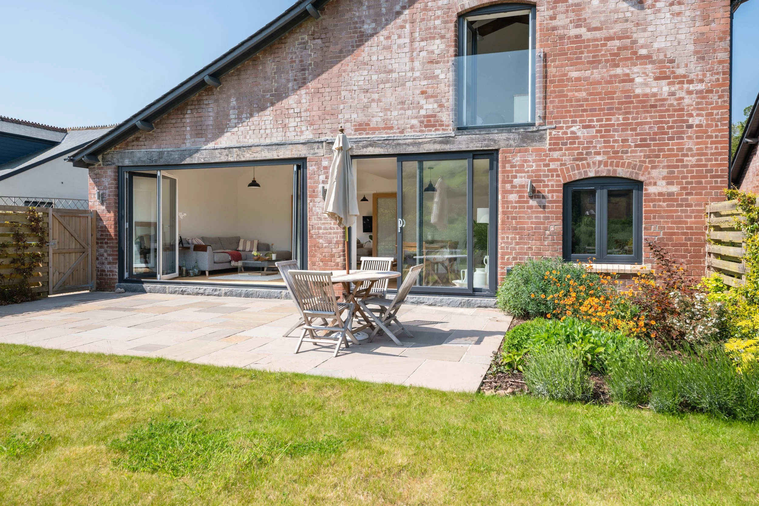 A brick house with large glass sliding doors opening to a patio with outdoor furniture. There is a flower bed with yellow and orange flowers and a green lawn.