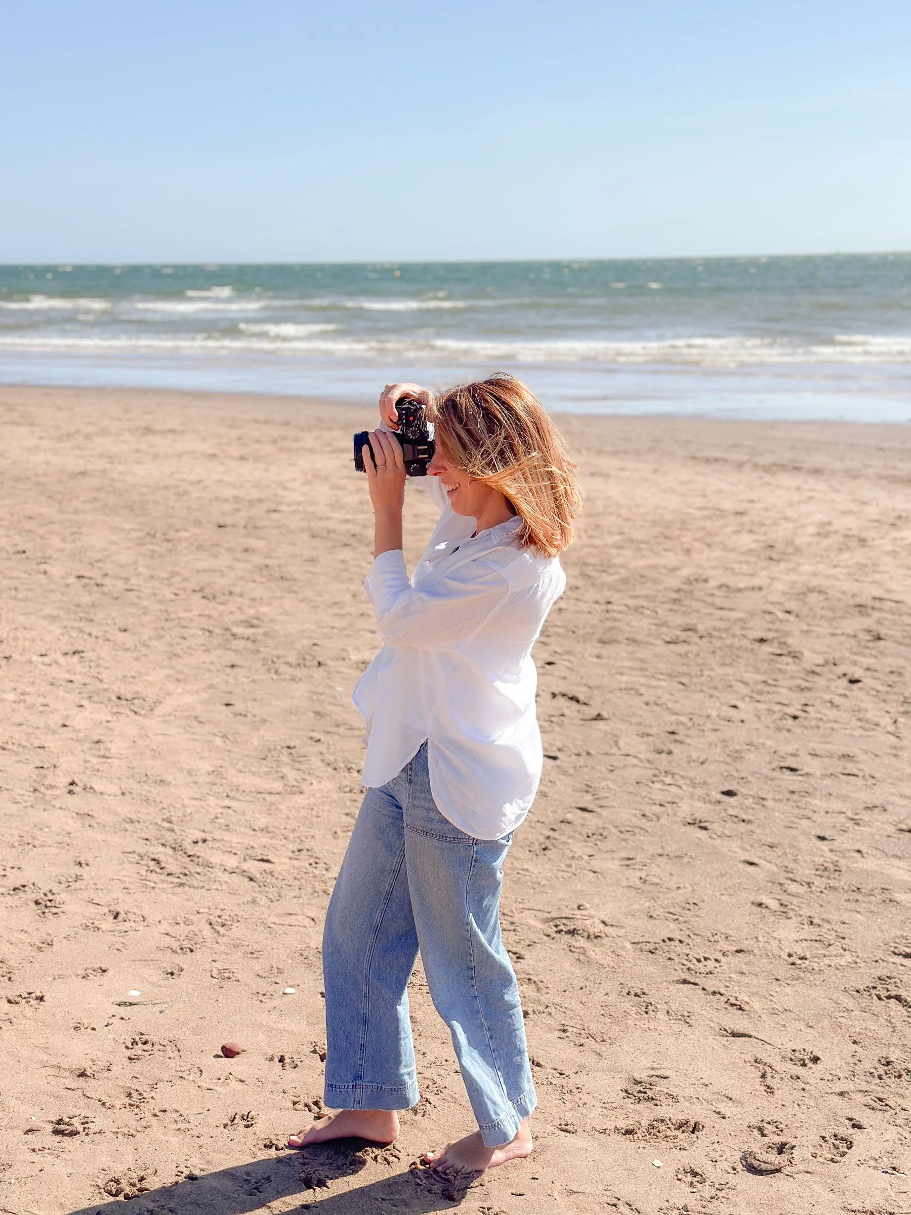 Woman standing on the beach taking a photo with a camera, wearing a white shirt and jeans, barefoot, with the ocean in the background.