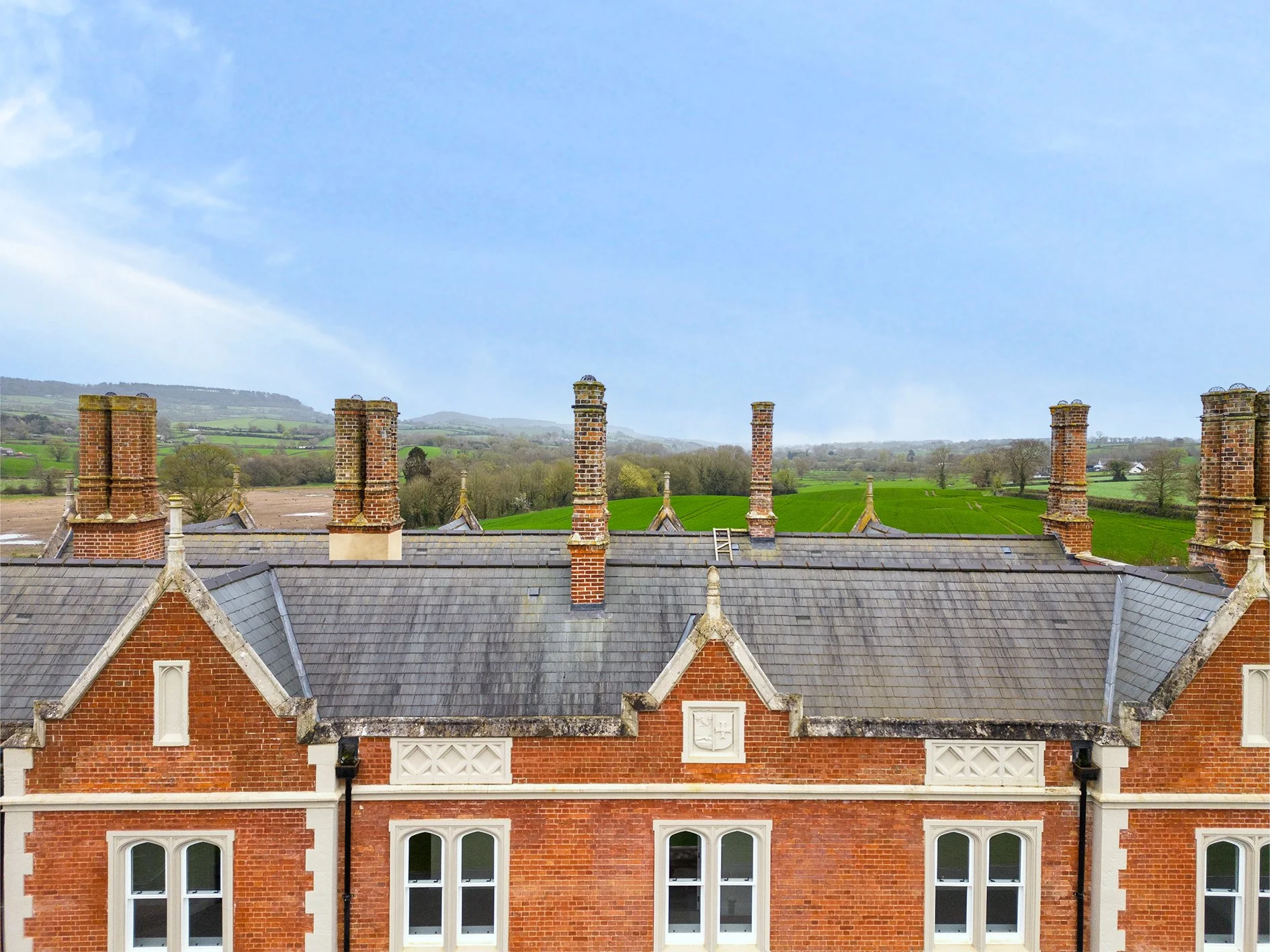 View of rooftops with brick chimneys, surrounding green fields, and distant hills under a clear blue sky.