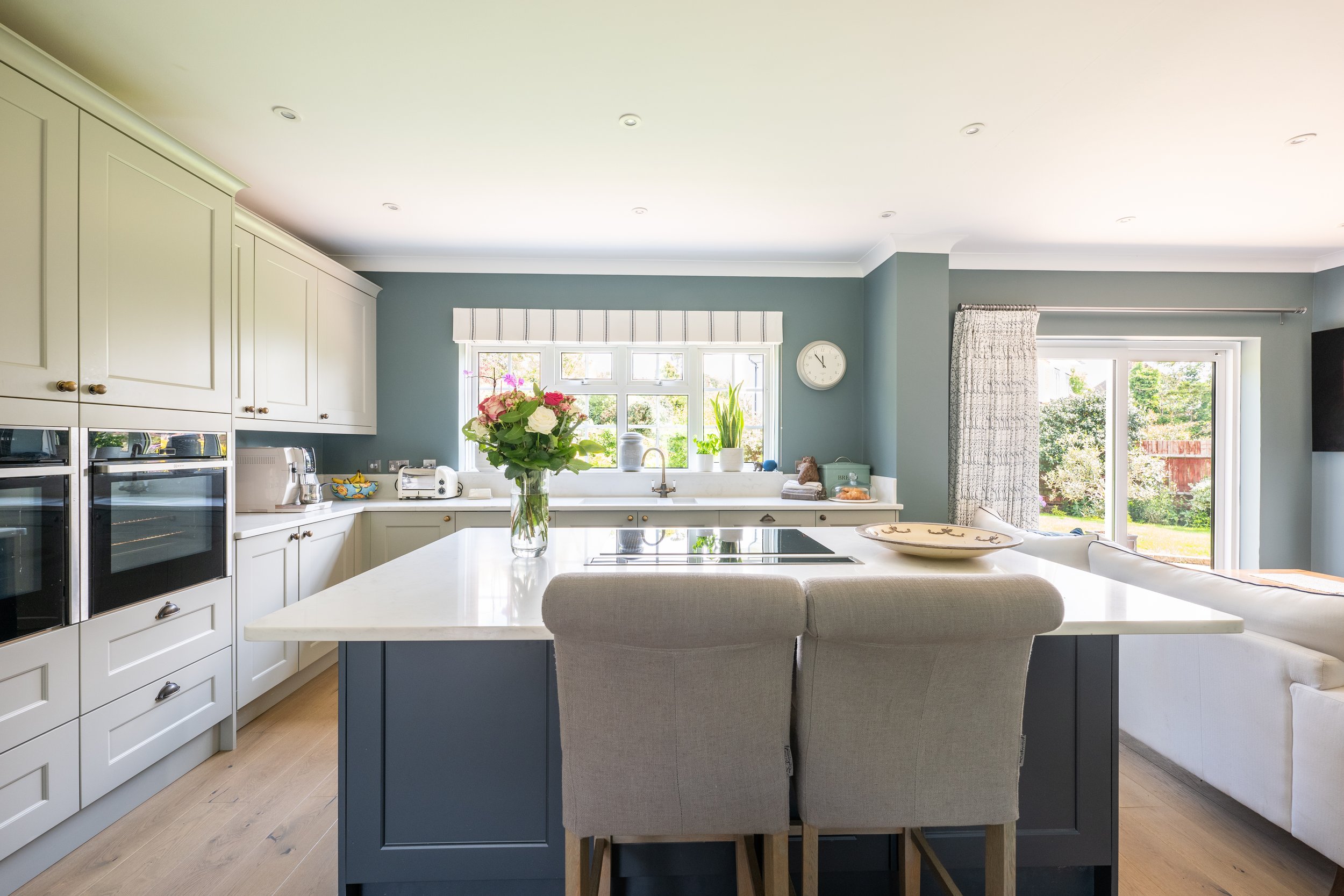 Bright kitchen with white cabinets, a blue island, and large windows with curtains, decorated with a vase of pink and white flowers on the island.