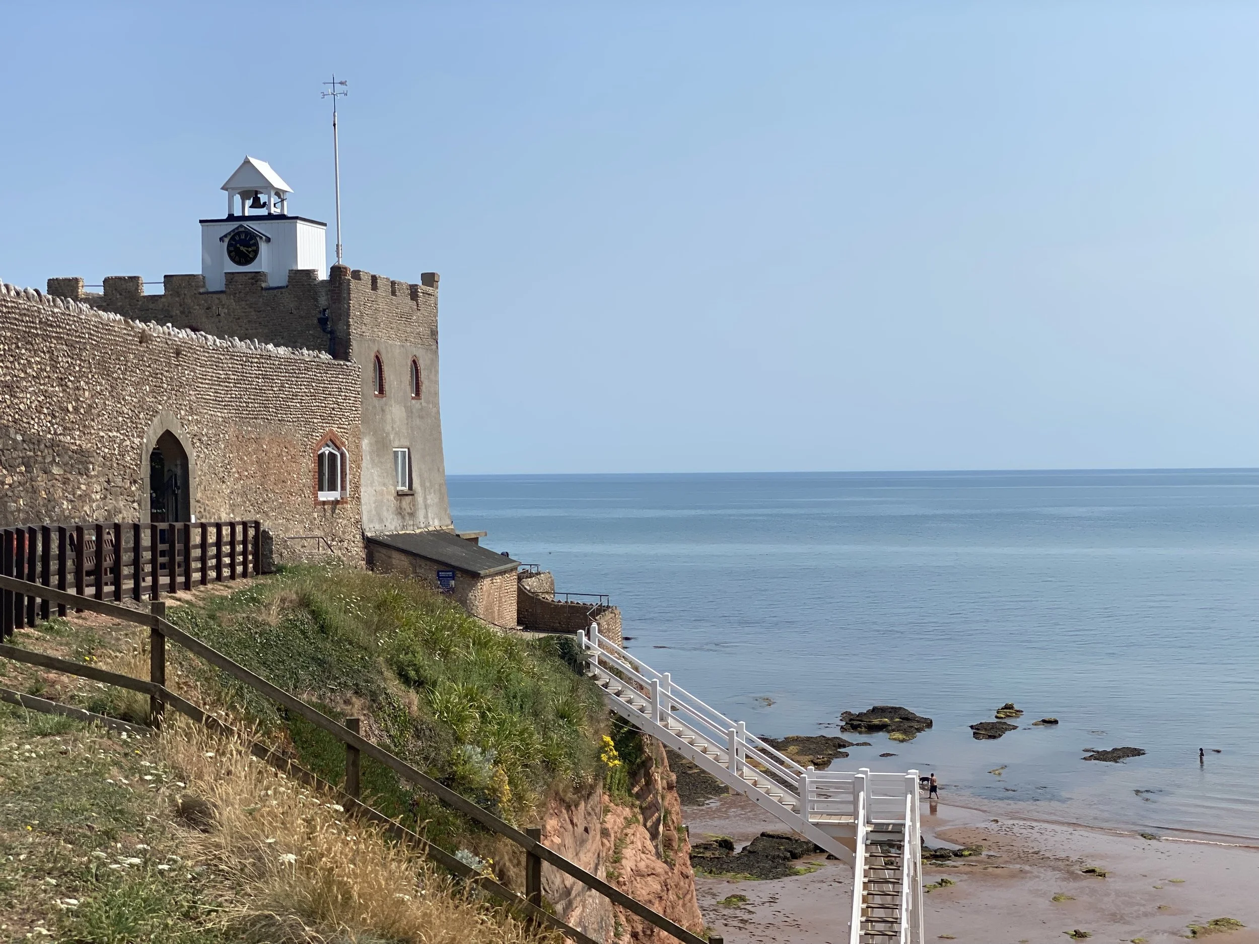 A seaside castle overlooking the ocean with a clock tower, stairs leading down to the beach, and a few people walking along the shore on a clear day.