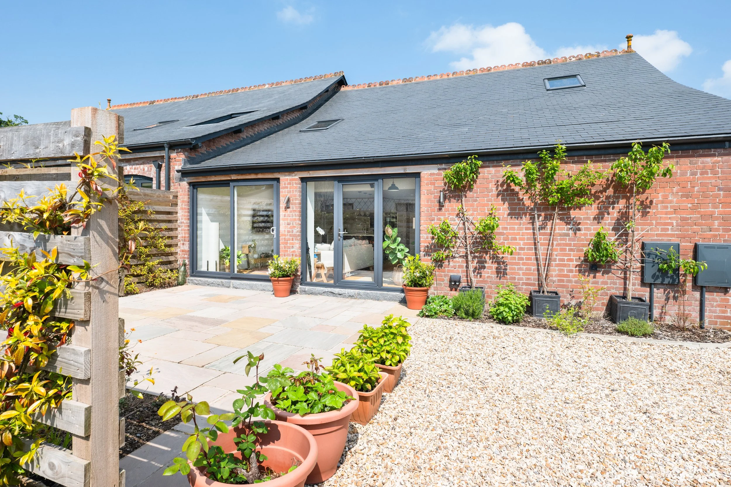 Backyard view of a brick house with sliding glass doors, potted plants, and a garden area with trees, plants, and a gravel pathway under a blue sky.