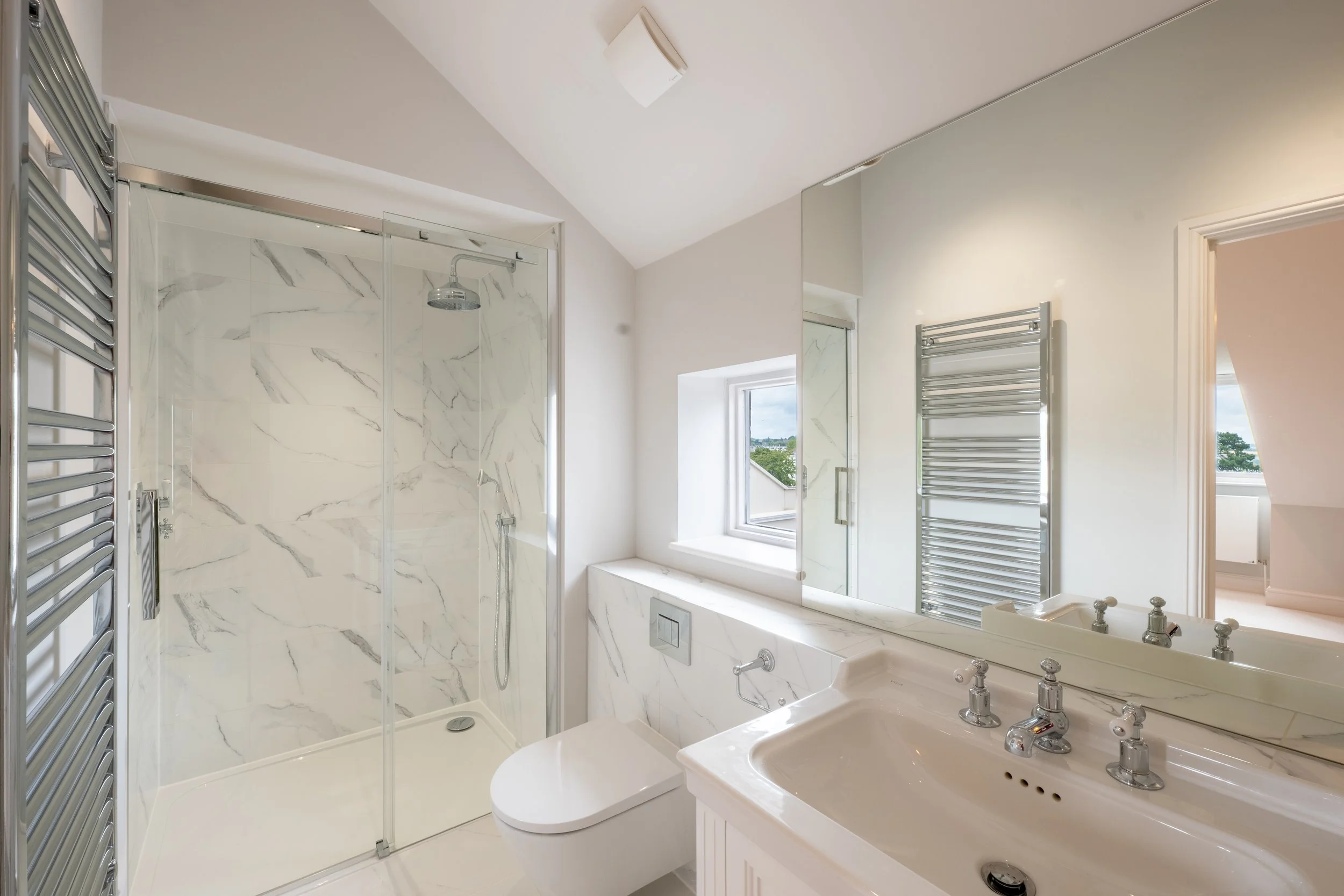A modern bathroom featuring a glass-enclosed shower with white marble walls, a window with a view of trees and sky, a white sink with chrome fixtures, a large mirror, a heated towel rail, and a white aesthetic.