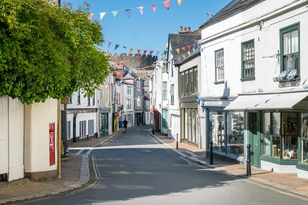 A quiet street in a small town with colorful bunting flags hanging overhead on a sunny day. The street is lined with white buildings with storefronts and windows, one of which appears to be an art gallery or shop. There is a large leafy green tree on