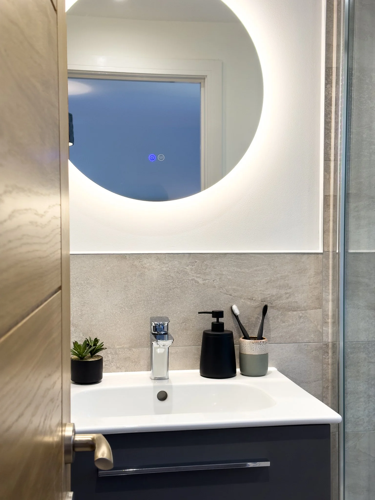 A modern bathroom countertop with a white sink, black and gray vases, a small green plant, and a mirror with built-in lighting.