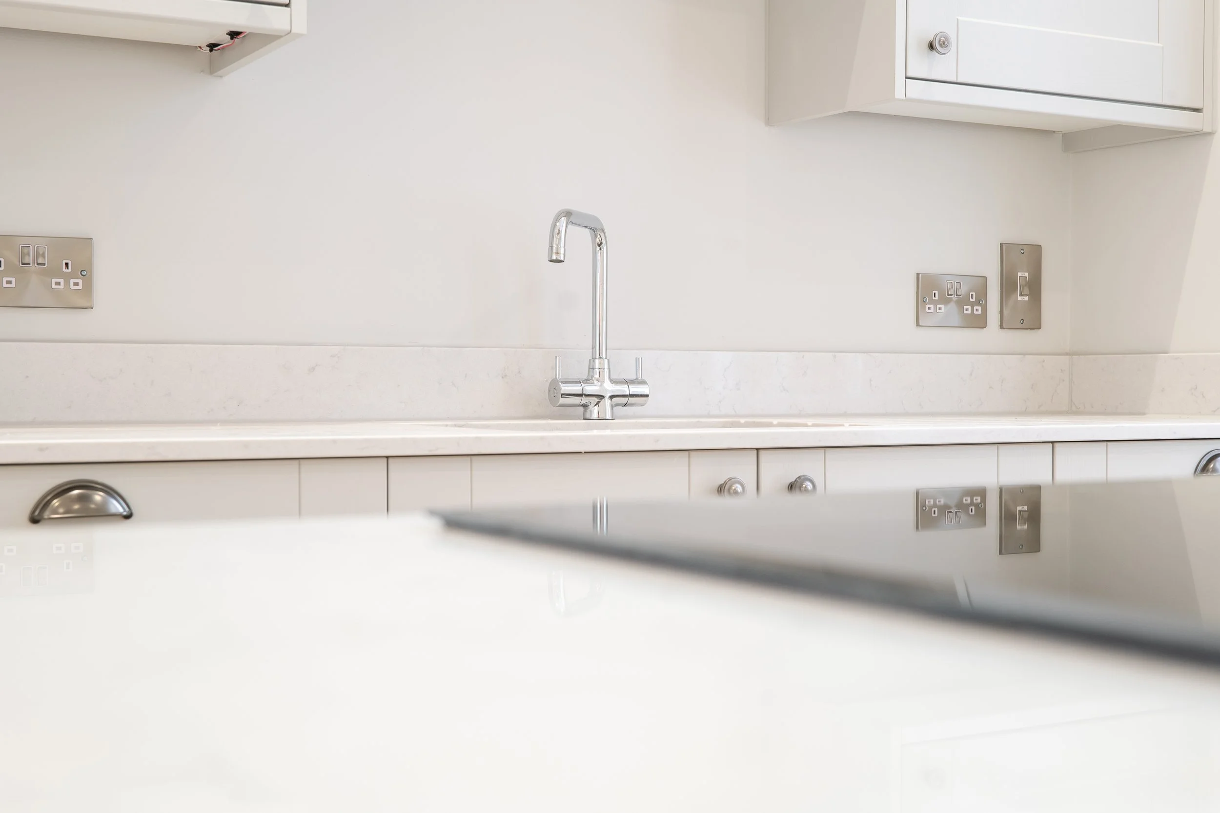 Kitchen sink with faucet, outlet sockets, and white cabinetry against a white wall.