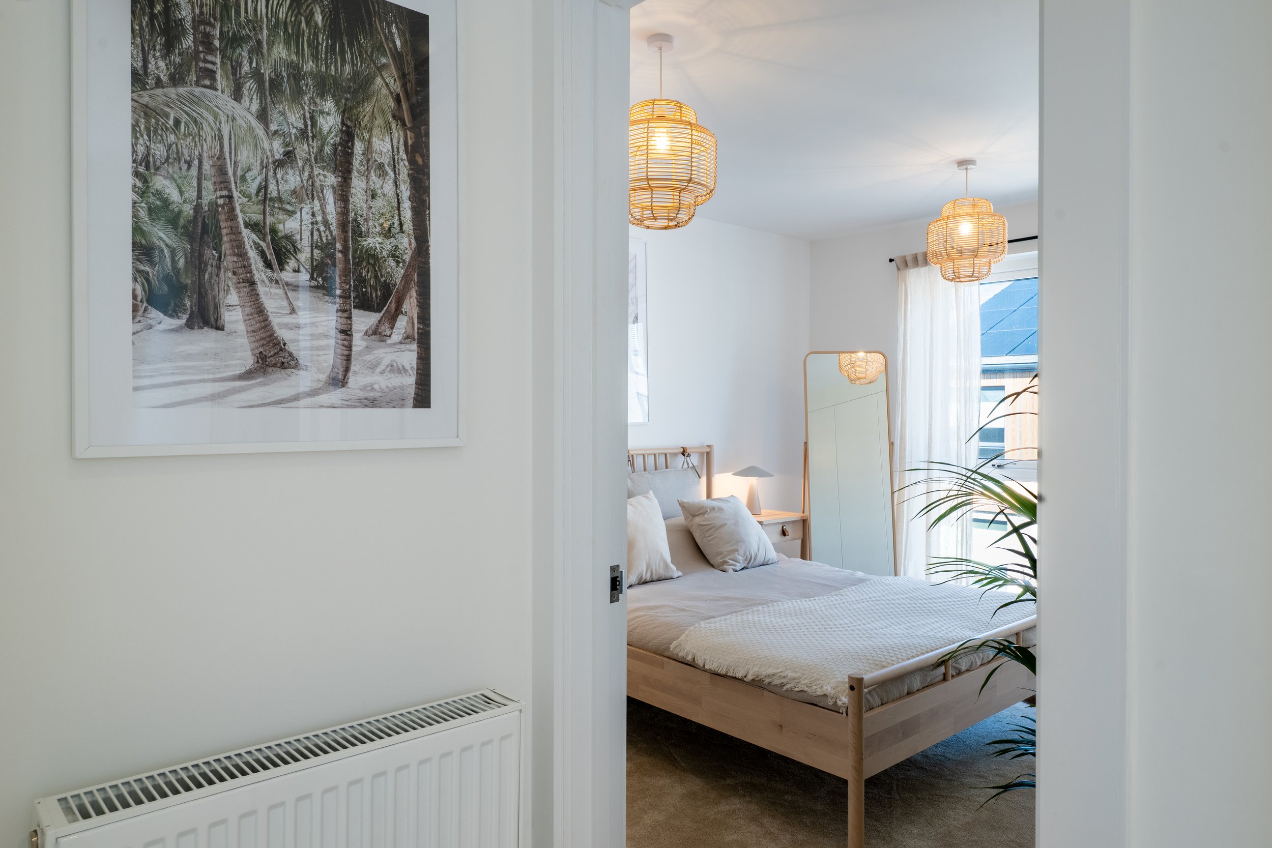 A view into a bedroom through a partially open white door, showing a wooden bed with white bedding and pillows, two hanging rattan pendant lights, a window with sheer off-white curtains, a standing mirror, and some indoor plants.