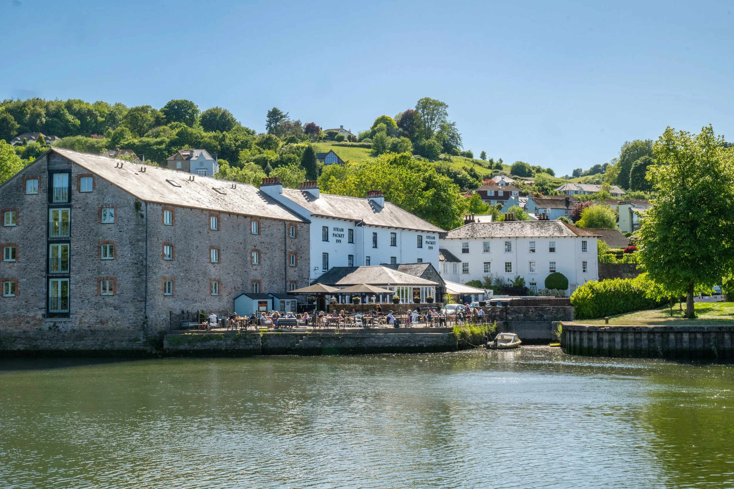 A scenic view of a river with green water in the foreground, and a riverside restaurant and buildings along the bank, with a lush green hill and homes in the background under a clear blue sky.