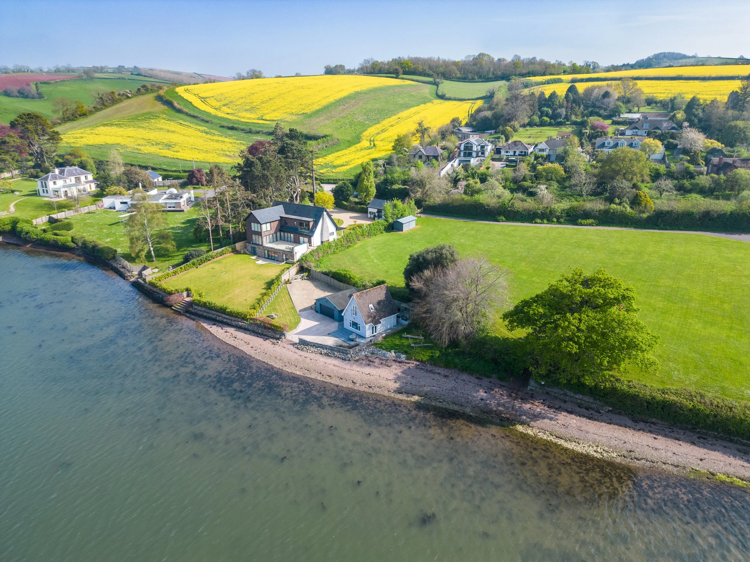 Aerial view of a coastal village with houses surrounded by green trees, fields, and rolling hills. There is a body of water in the foreground with a shoreline.