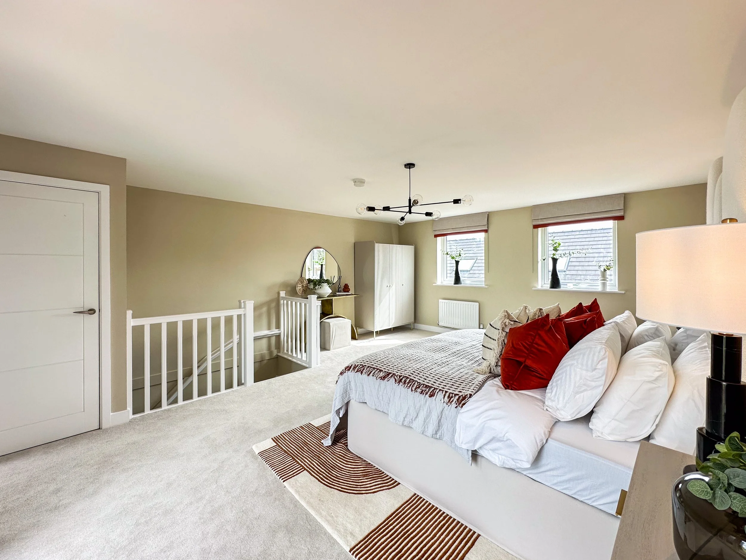 Bright living room with a white sofa, red pillows, a blanket, a black side table, and houseplants. Large windows with beige blinds let in natural light. A modern black chandelier hangs from the ceiling.