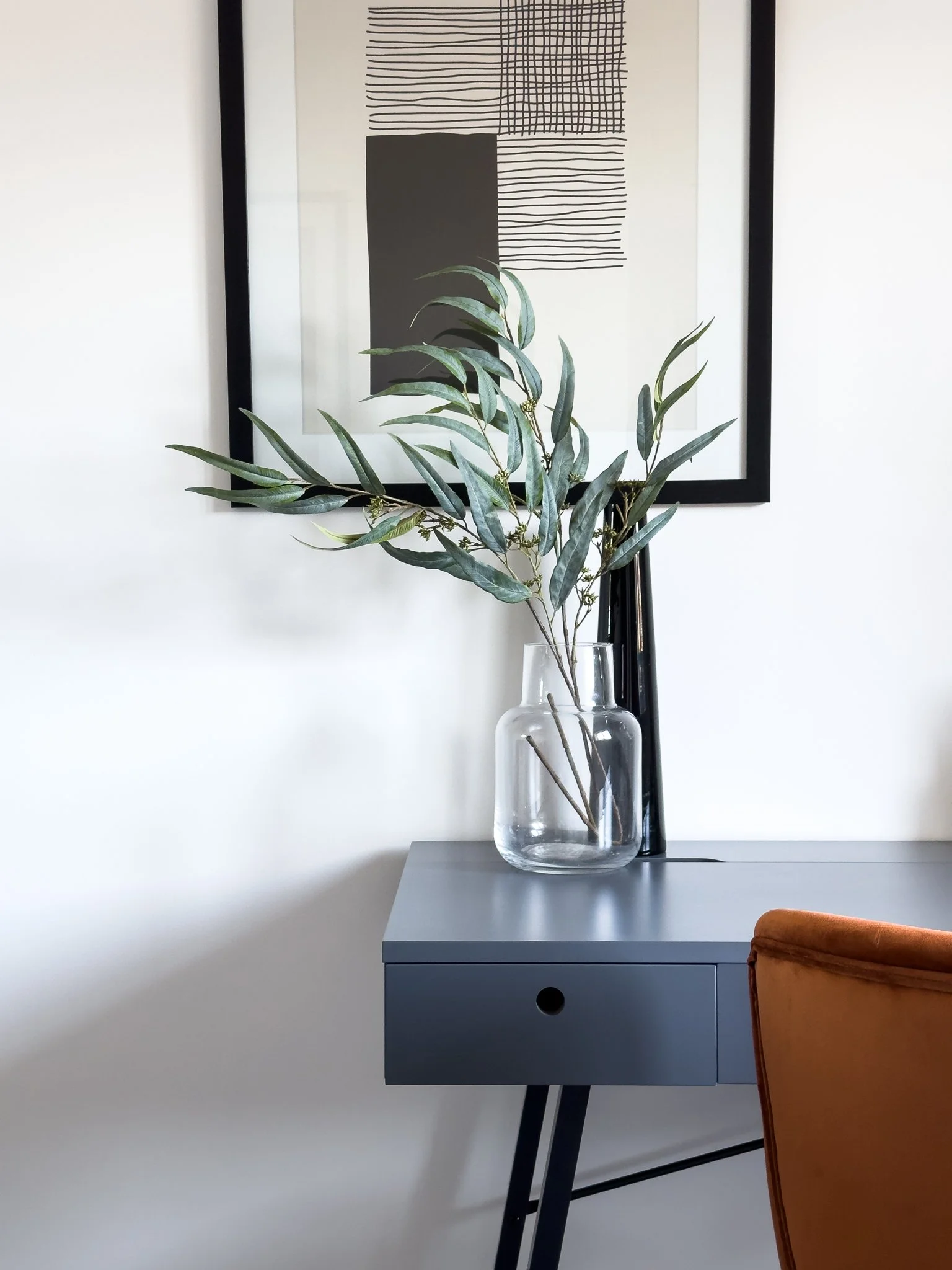 A minimalist interior scene with a black-framed abstract art piece hanging on a white wall, a transparent glass vase with green leafy branches on a gray side table, and an orange chair partially visible at the bottom right corner.