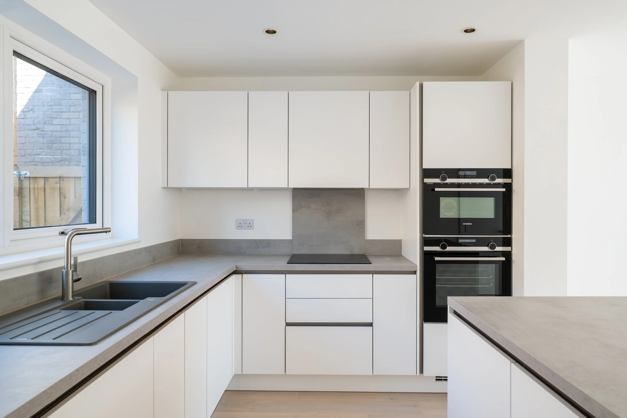 Modern kitchen with white cabinets, black double oven, window with a view of a brick wall and wooden fence, sink with faucet, and beige countertops.
