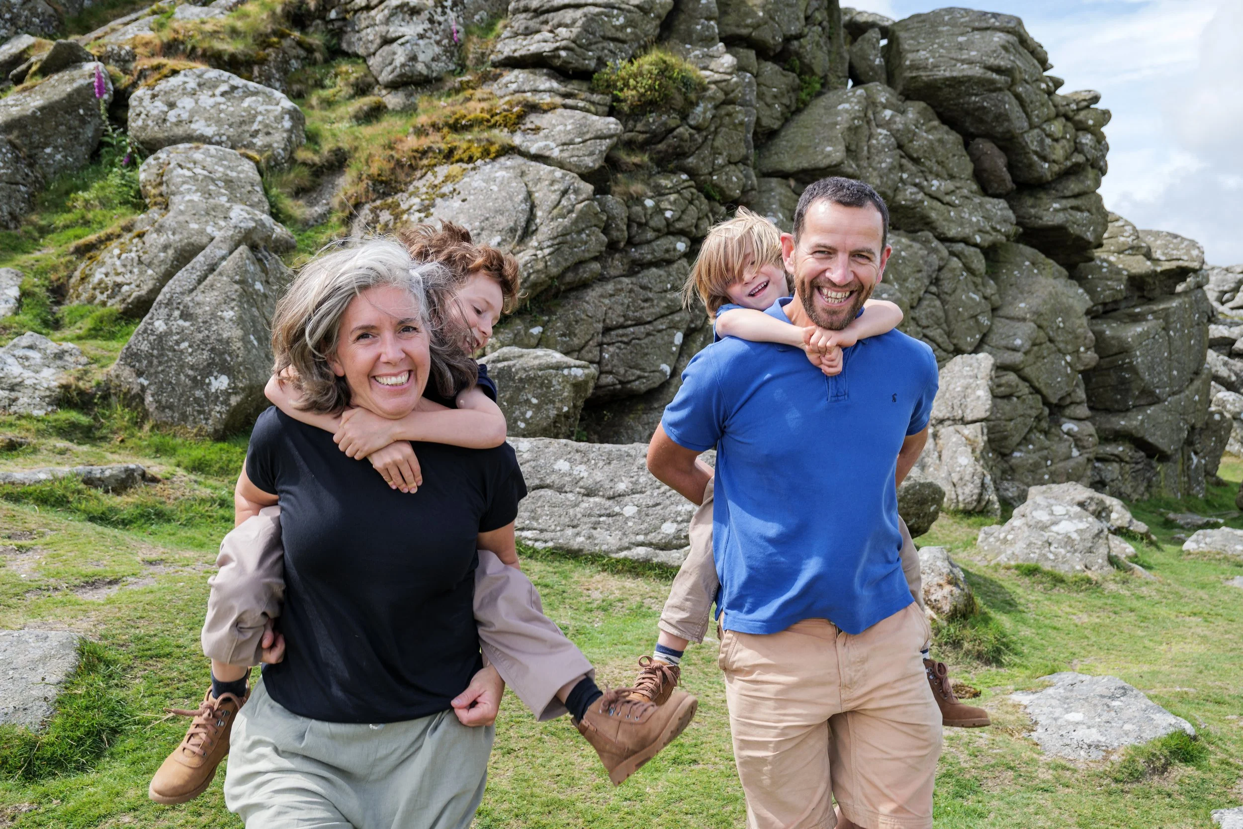 Family of four hiking outdoors near rocky terrain, with two children on their parents' shoulders, smiling.