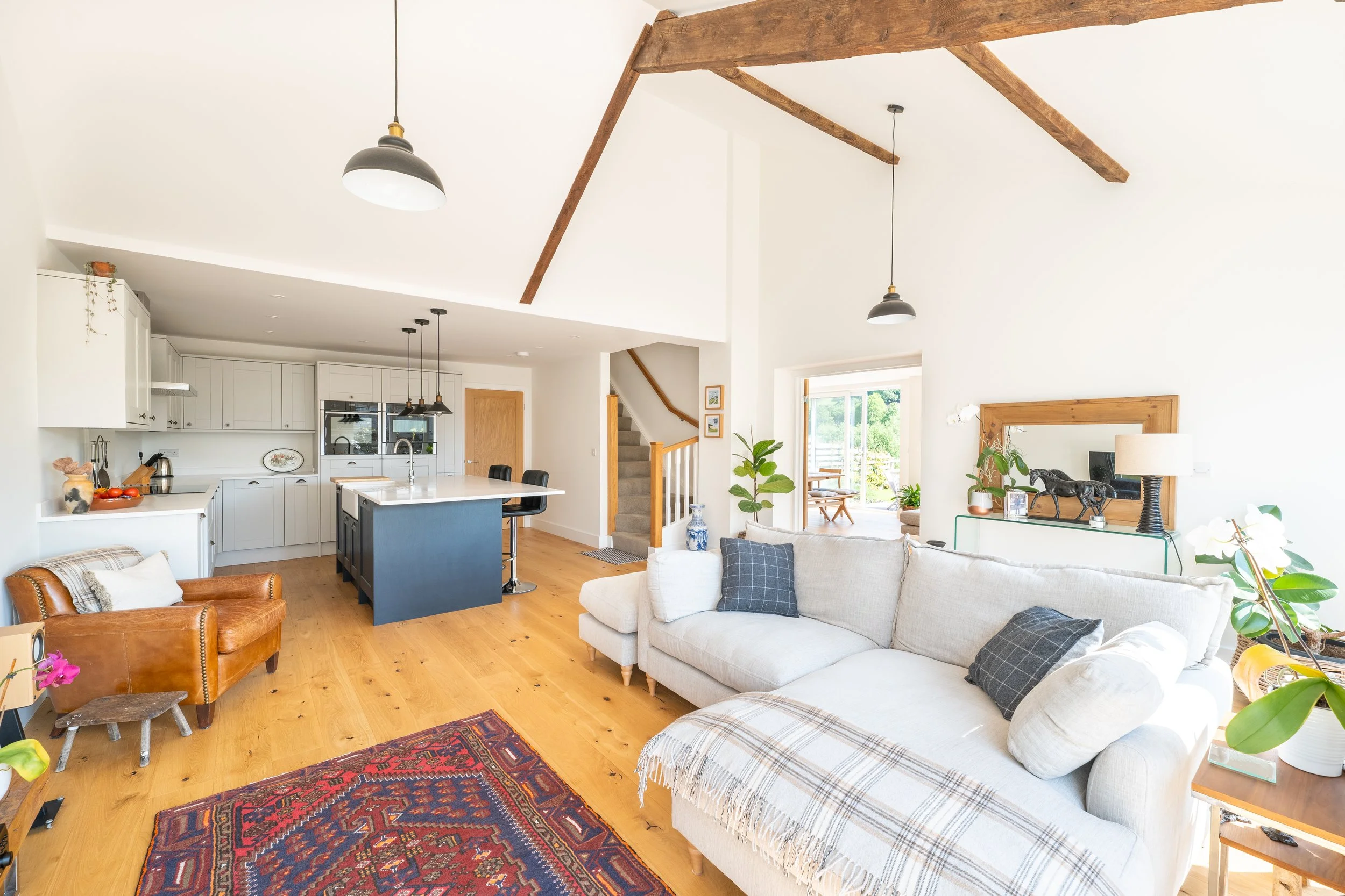 A bright, open-concept living space with a white sofa and decorative pillows, a wooden armchair, a colorful rug, a kitchen with white cabinets, a blue kitchen island, pendant lights, a staircase, and large windows with a view of greenery outside.