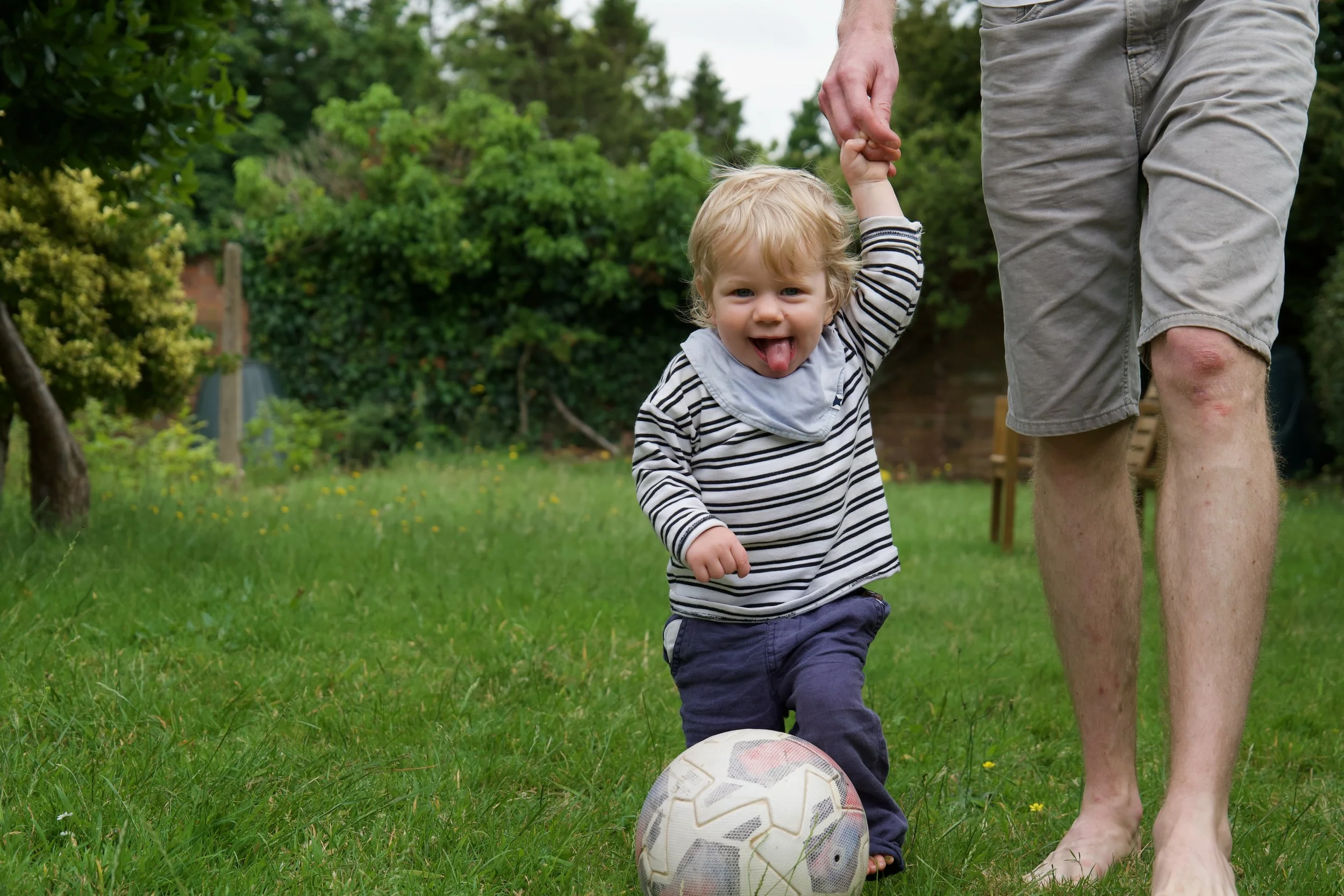 A young boy with blond hair and a striped shirt playing soccer outdoors, holding hands with an adult, on a grassy field with trees in the background.