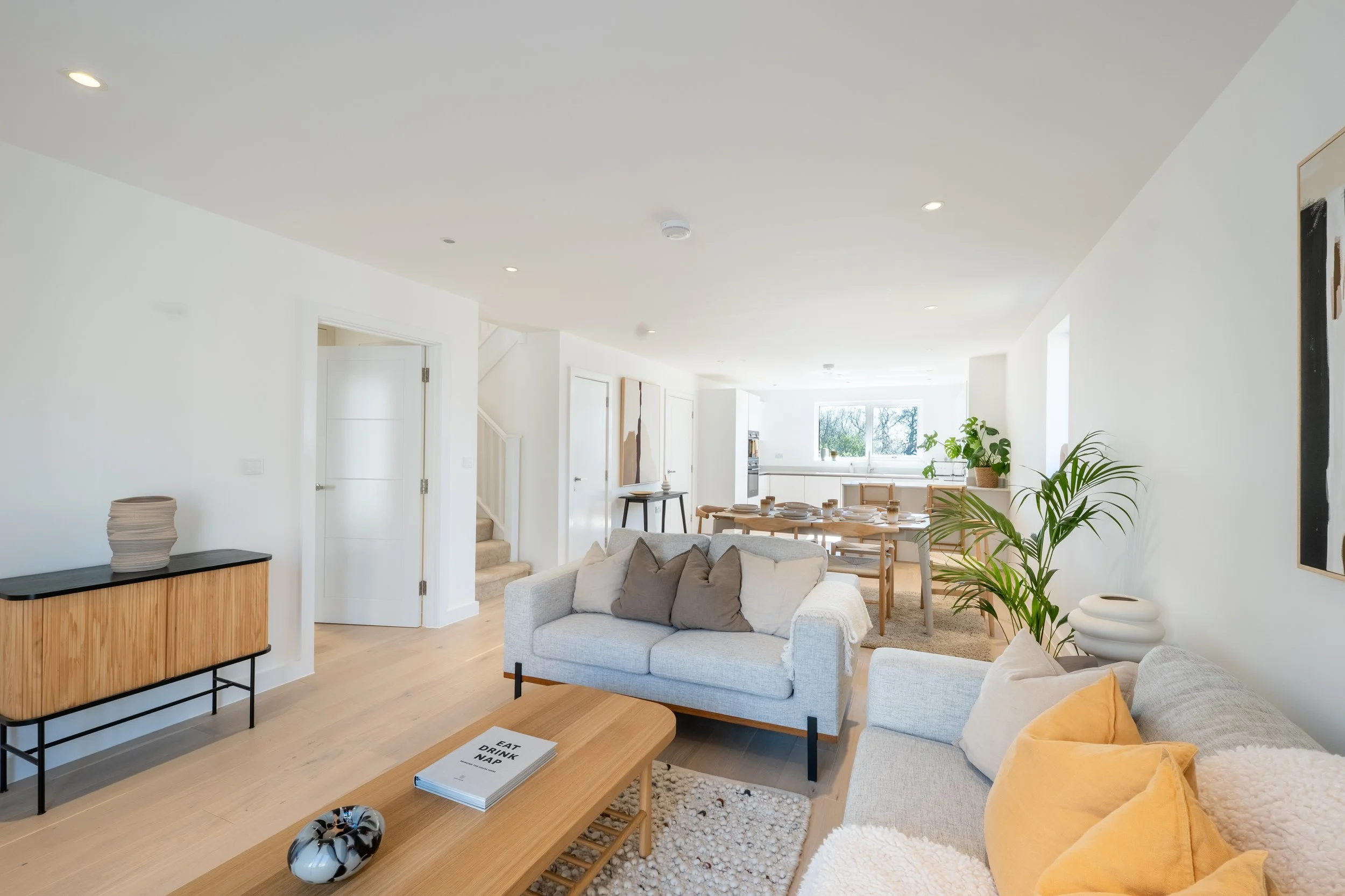 Open-concept modern living room with white walls, light wood flooring, gray sofas with pillows, a wooden coffee table with a book, potted plants, and a white kitchen in the background with large window.