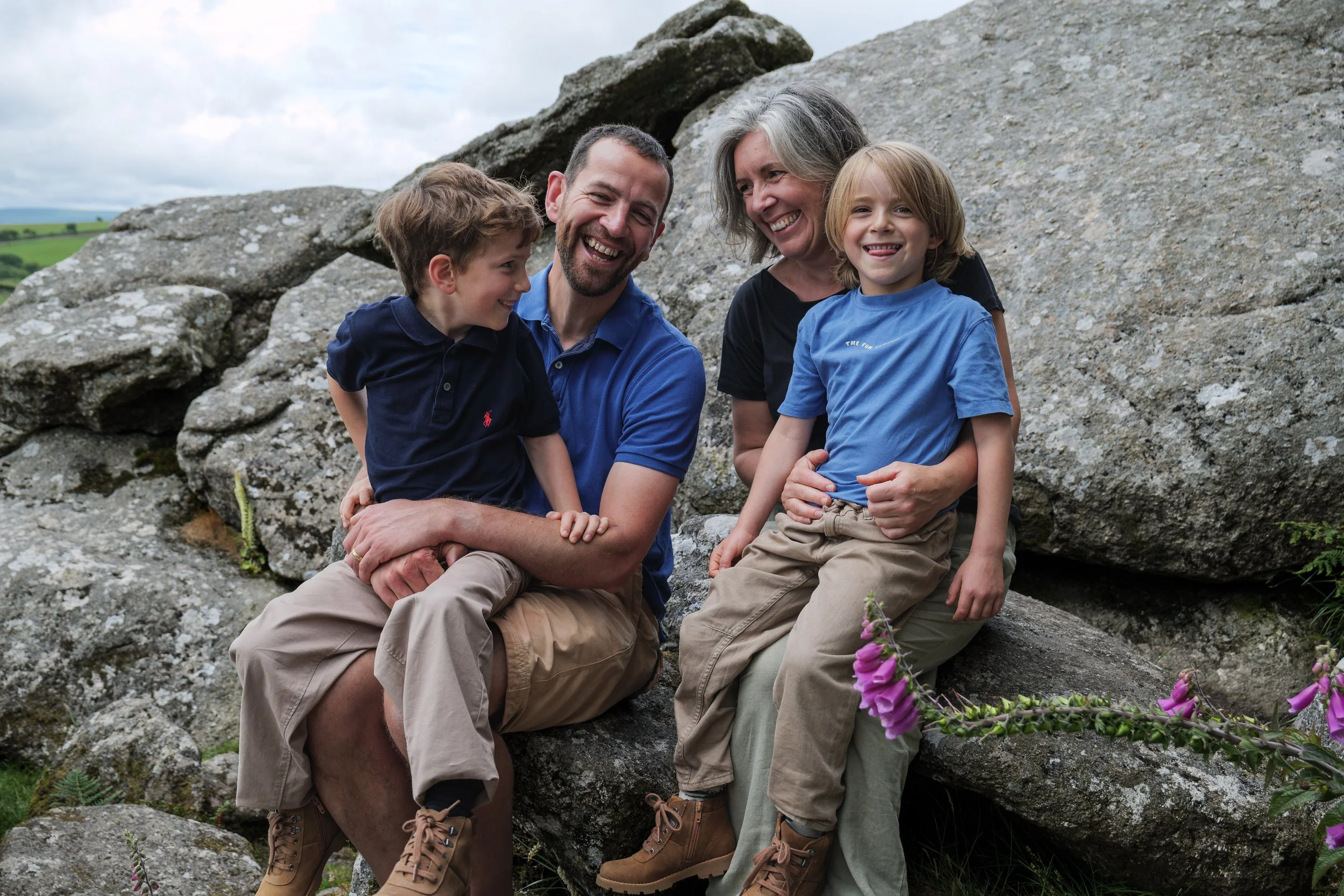 A family of four, including two children and two adults, sitting on rocks outdoors near flowering plants with a cloudy sky and green landscape in the background. They are smiling and appear to be enjoying nature.