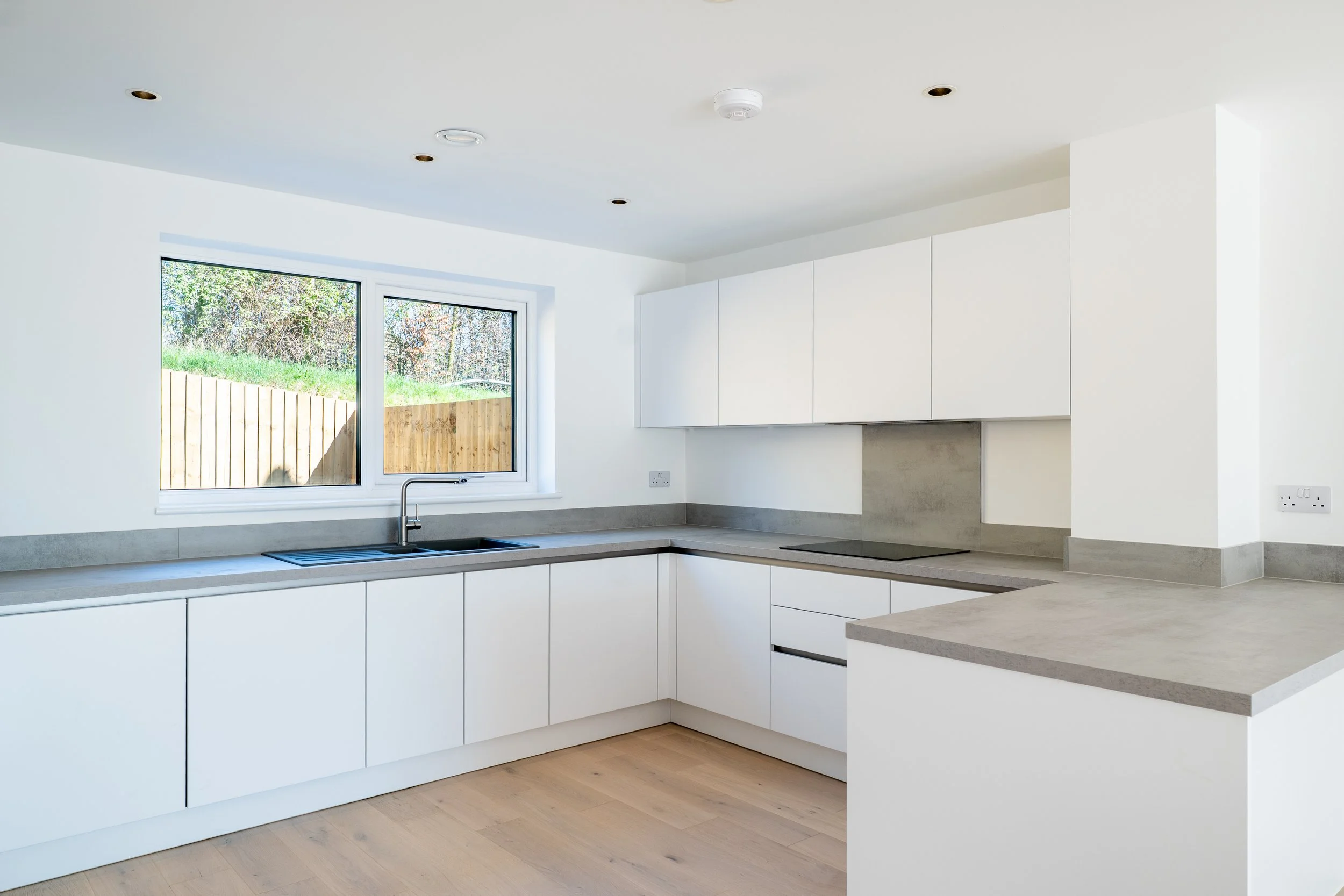 Modern kitchen with white cabinets, gray countertops, a window showing outdoor greenery, and a built-in stovetop.