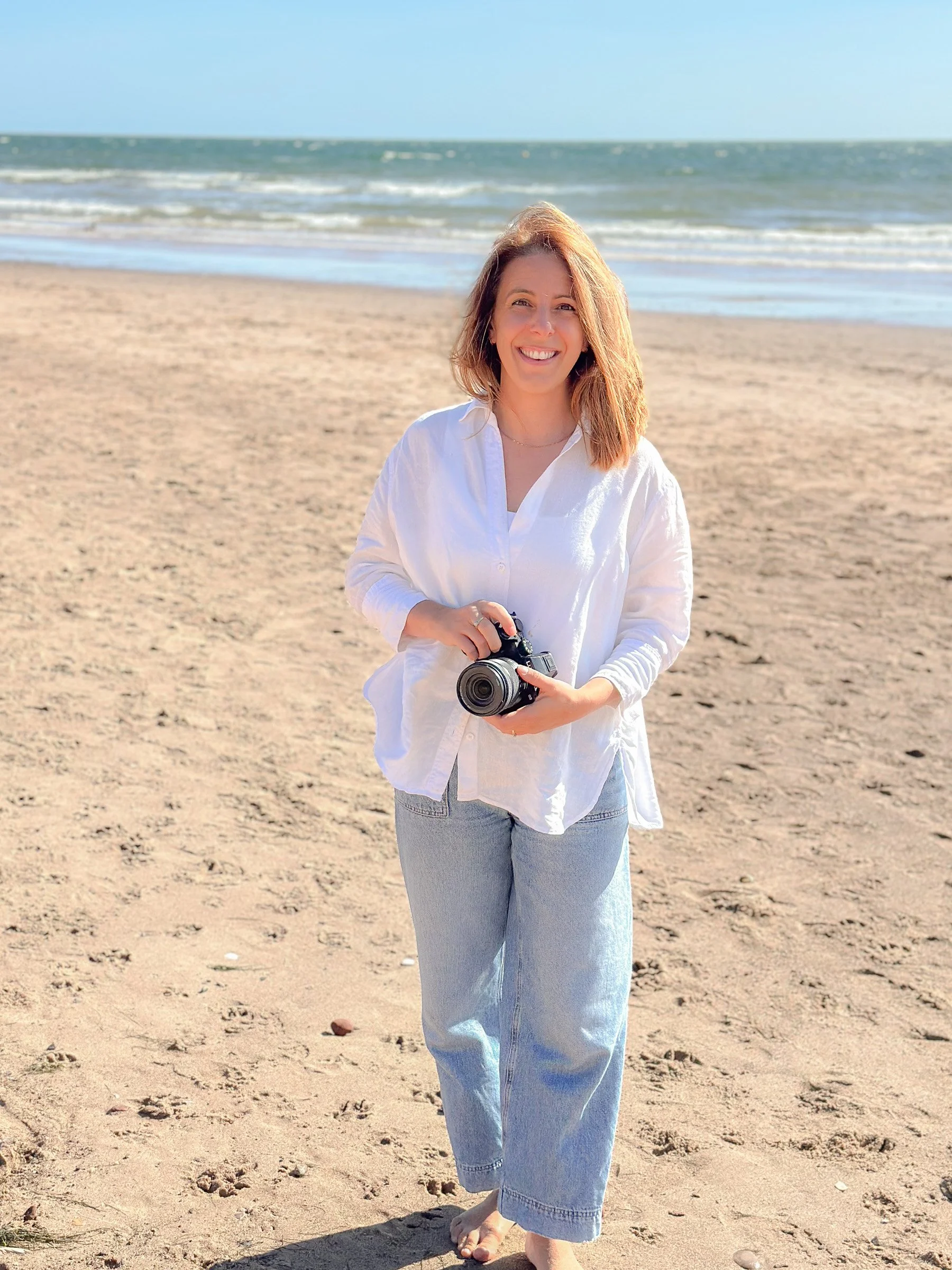 A woman with shoulder-length hair smiling, holding a camera, wearing a white blouse and light blue jeans, standing barefoot on a sandy beach with the ocean and clear blue sky in the background.