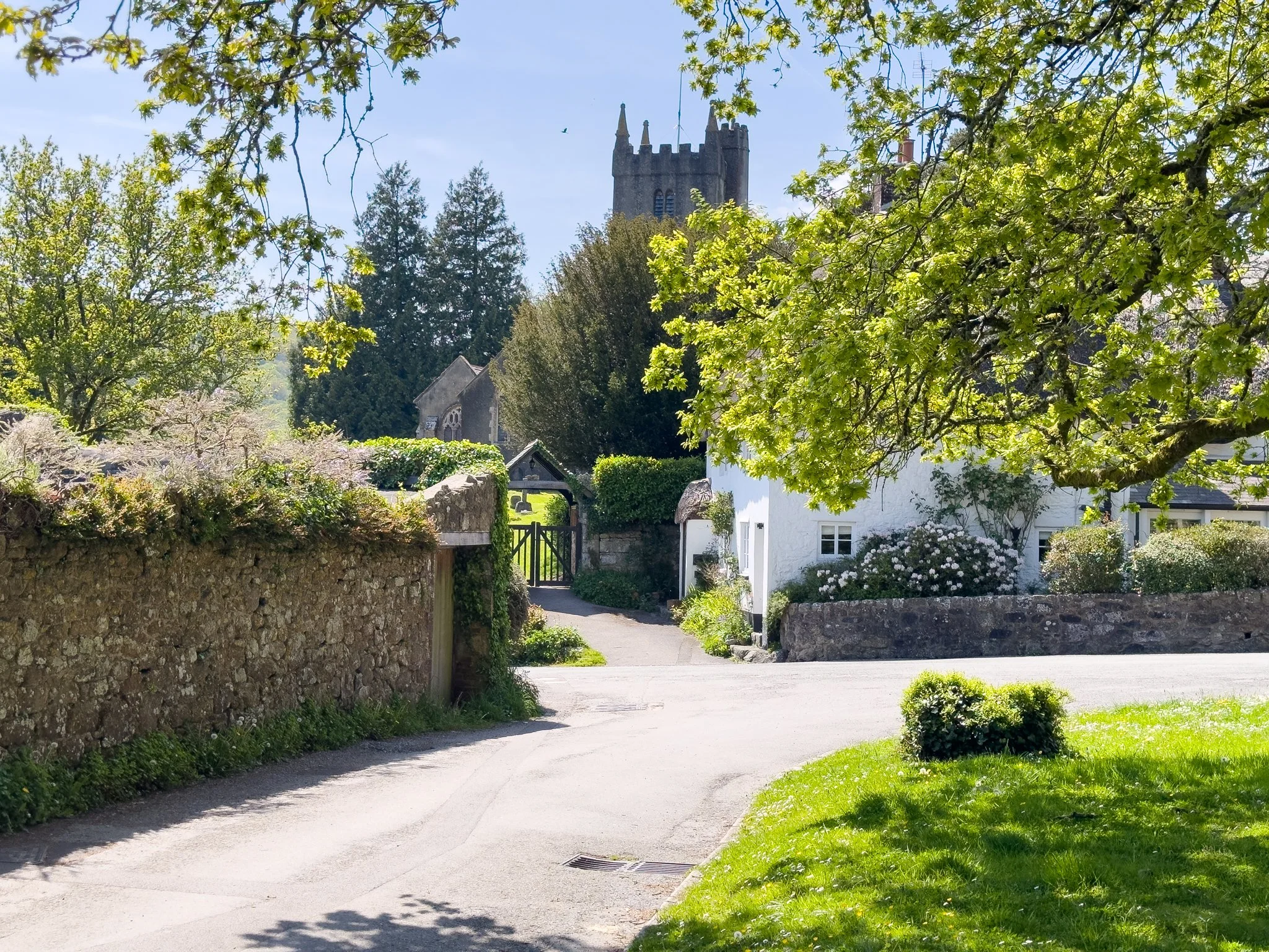 A peaceful village scene with stone walls, a church tower with pointed spires, surrounded by lush green trees and a clear blue sky.