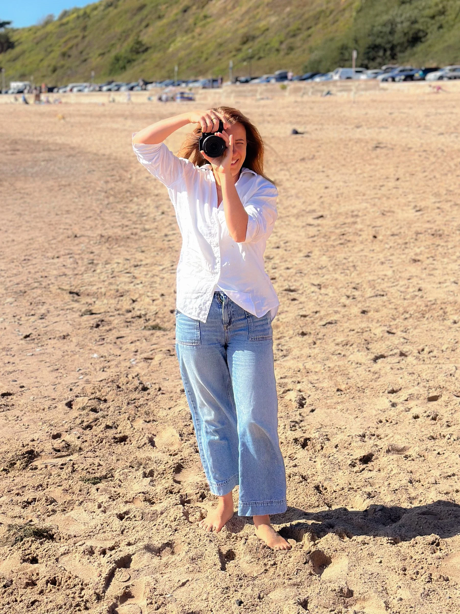 A woman standing barefoot on sandy beach, holding a camera to her face, with a hill and parked cars in the background on a sunny day.