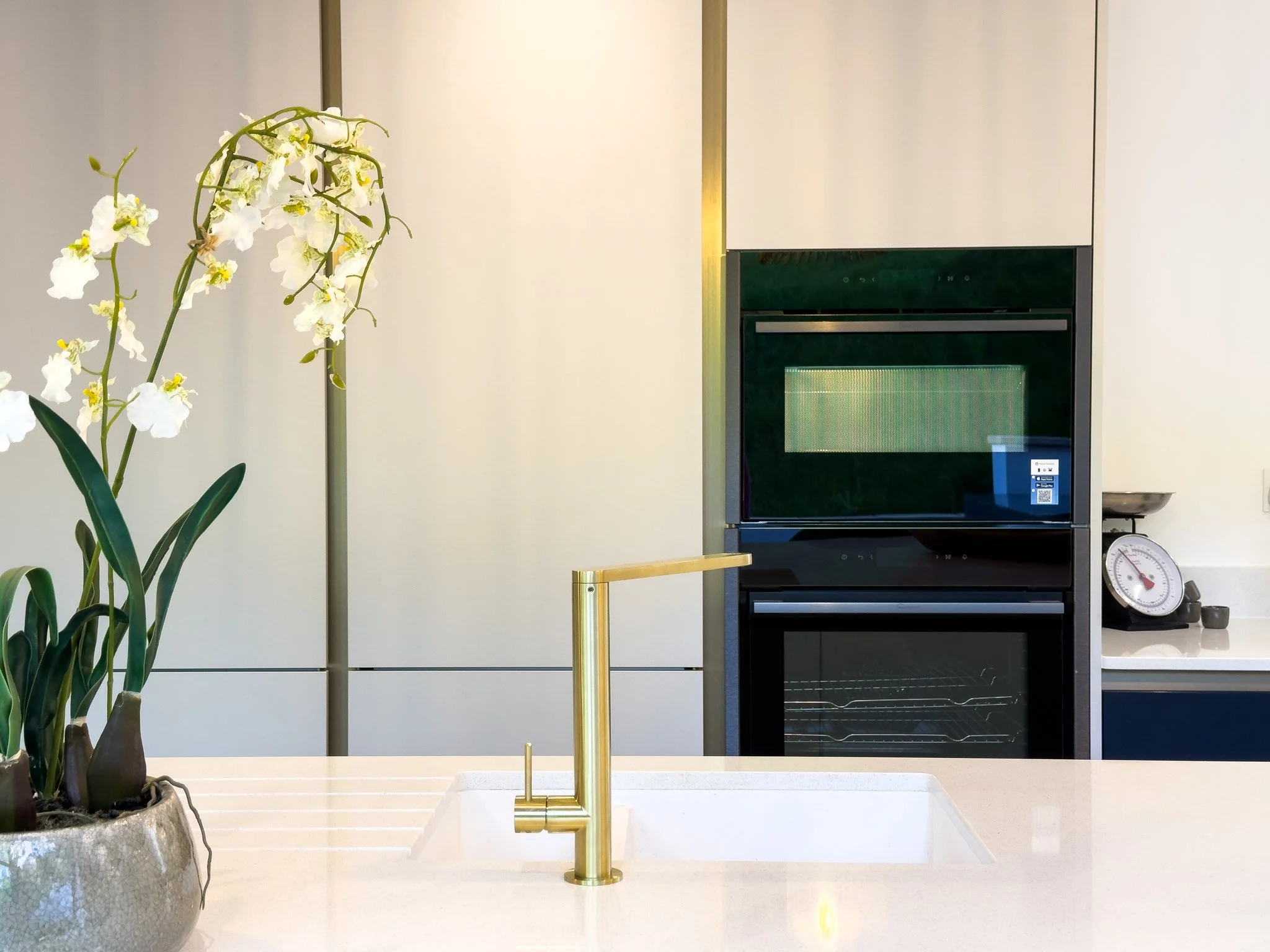 A modern kitchen with a white island sink, gold faucet, potted plant, and a built-in oven in the background.
