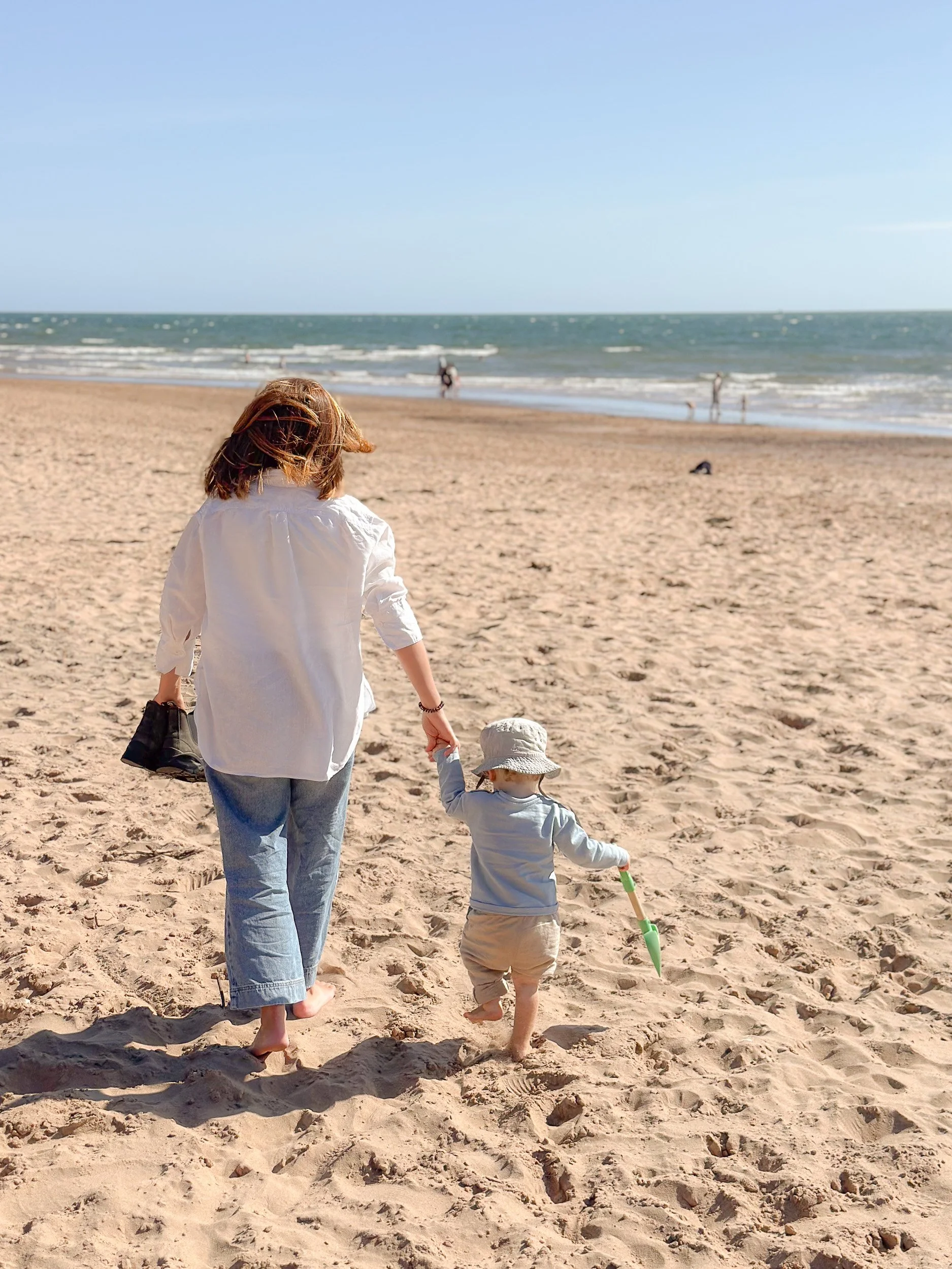 A woman and a young child walking on a sandy beach toward the ocean, with other people in the background near the water under a clear blue sky.