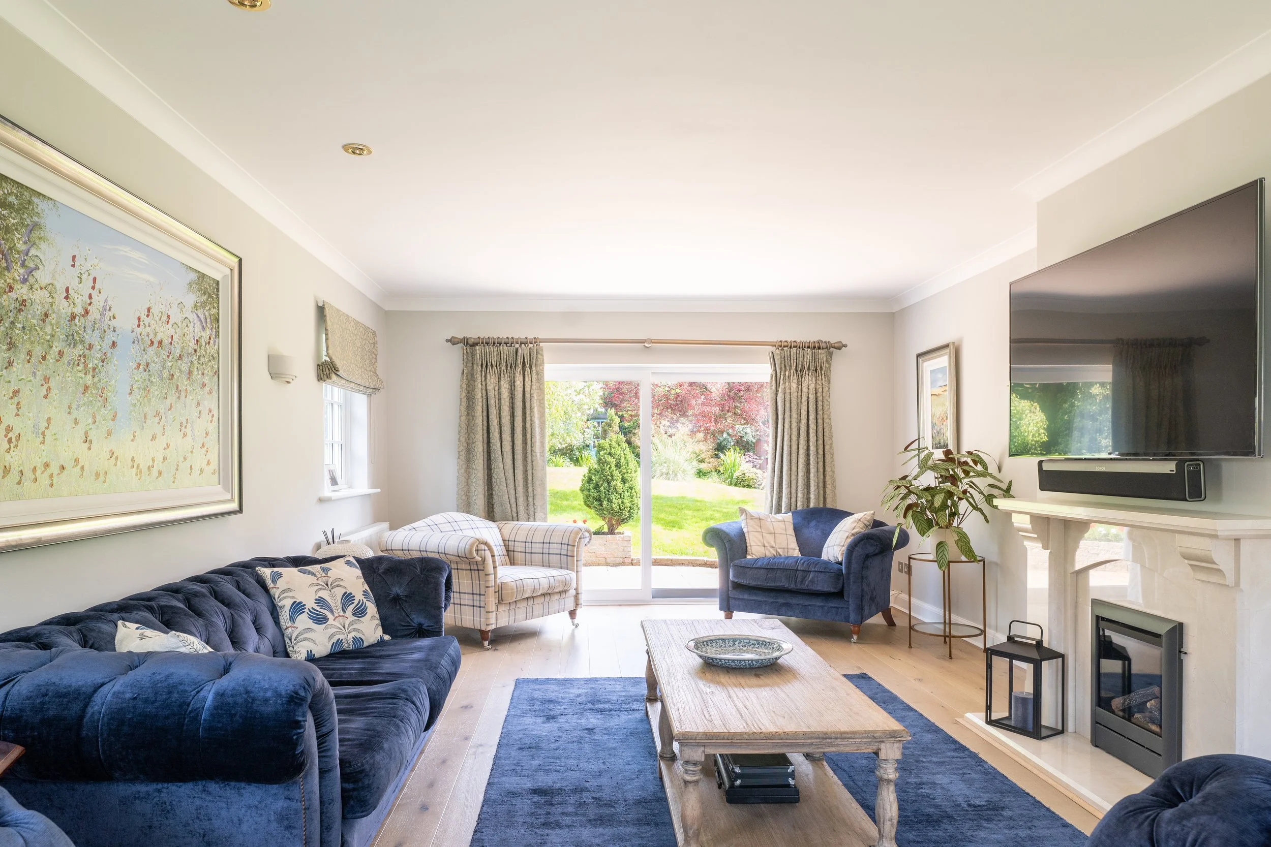Living room with navy blue velvet sofa and armchair, wooden coffee table, large window with curtains showing garden outside, wall art, wall-mounted TV, fireplace, potted plant, and hardwood floors.