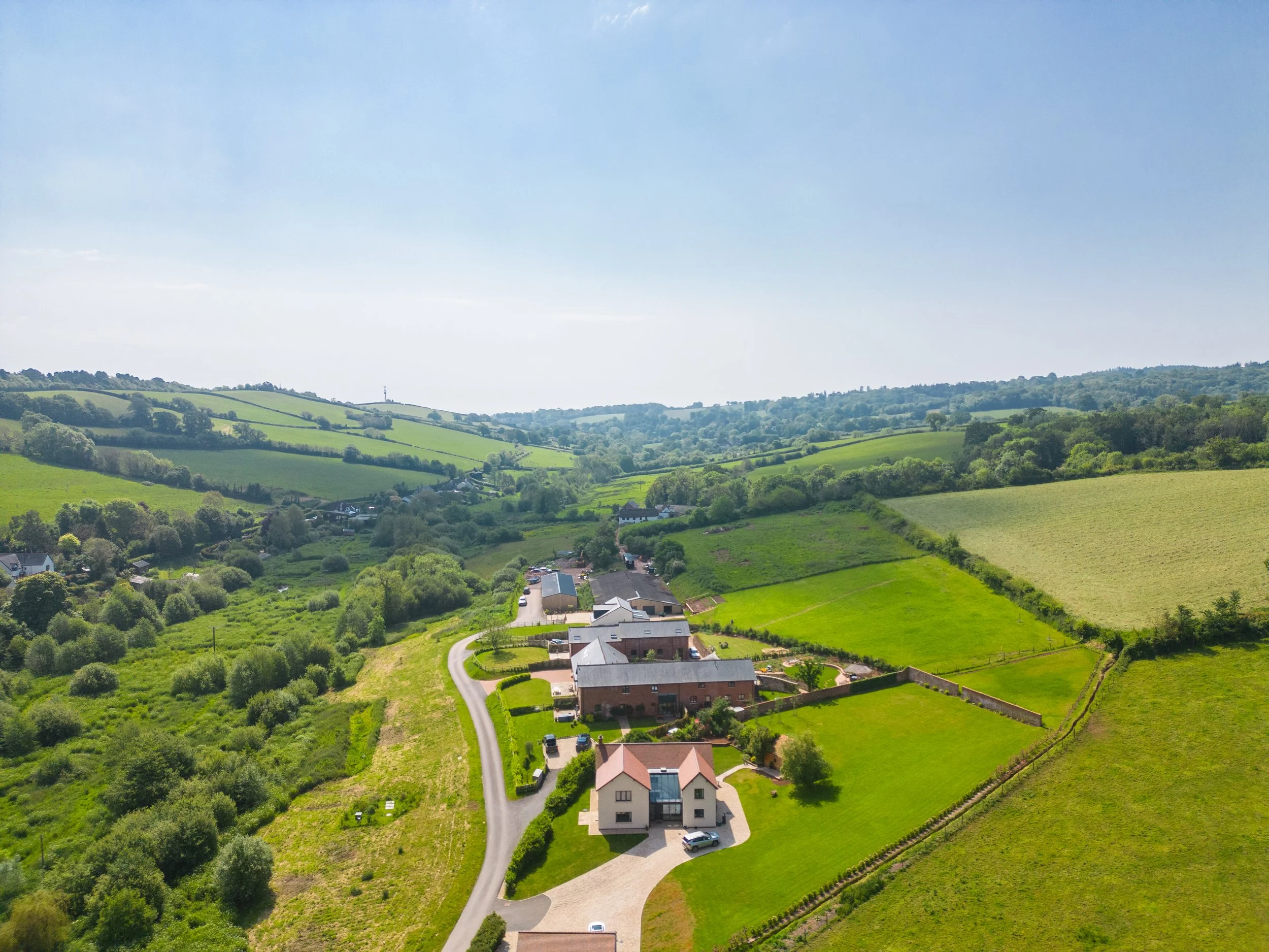 Aerial view of a rural landscape with rolling green hills, scattered houses, and a winding road leading to a modern white house with a driveway and parked cars.