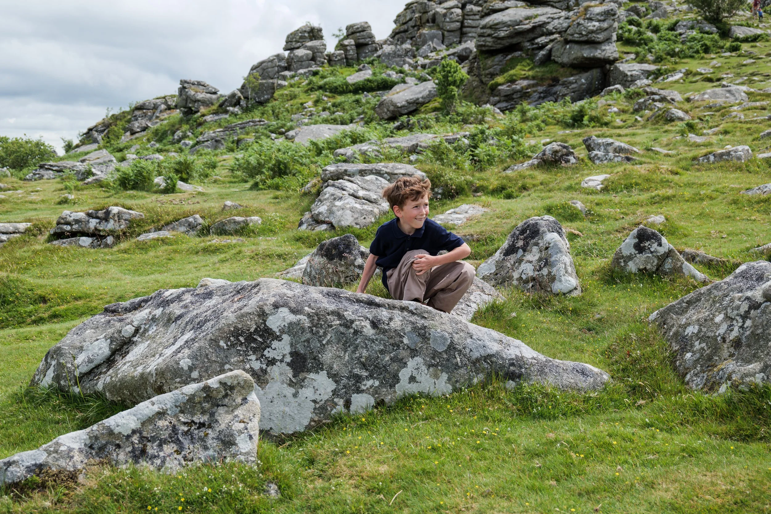 A young boy crouching behind a large rock in a grassy, rocky hillside with scattered bushes and trees under a cloudy sky.