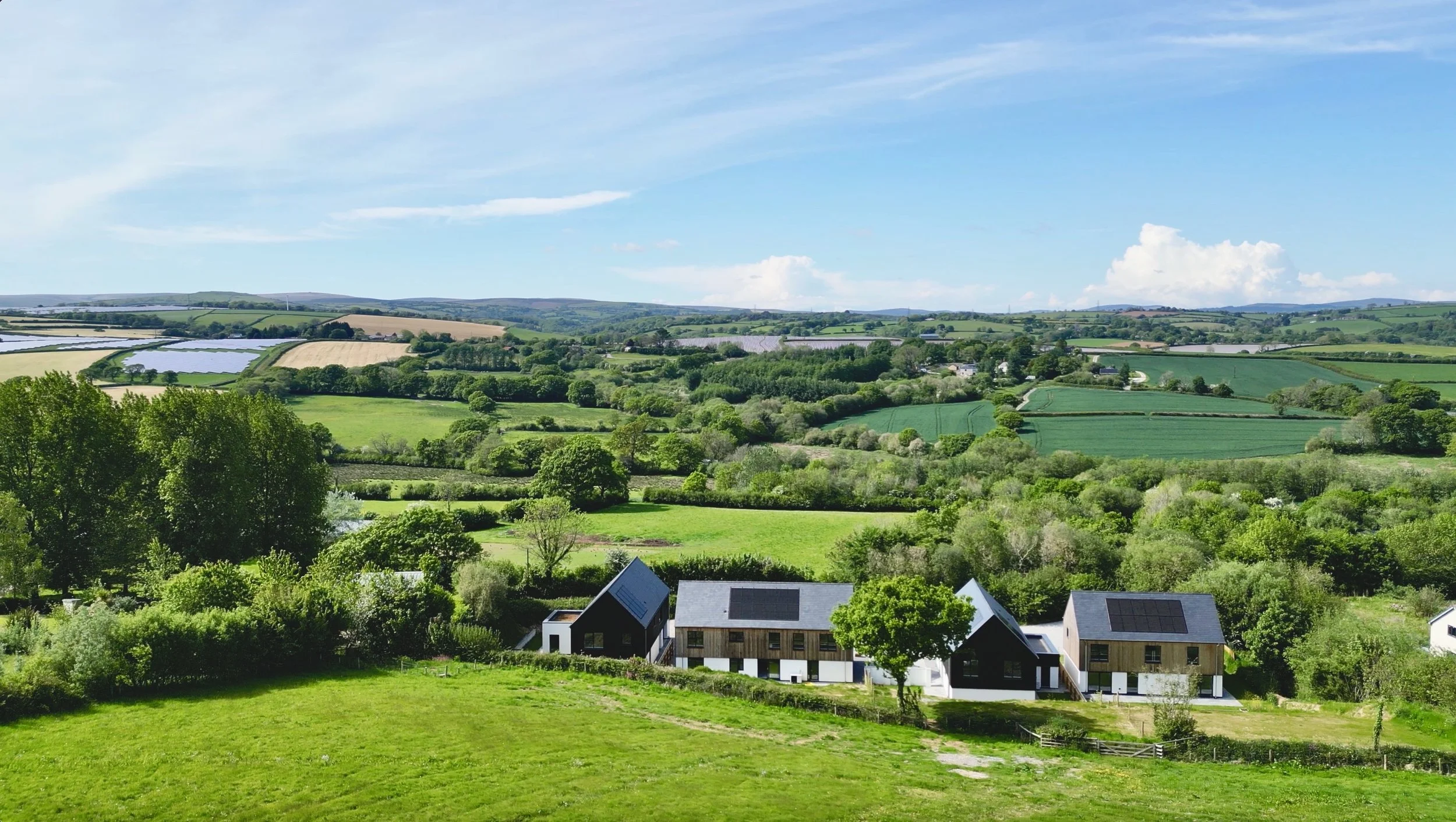Scenic view of a lush green valley with modern houses, rolling fields, and dense trees under a blue sky with some clouds.