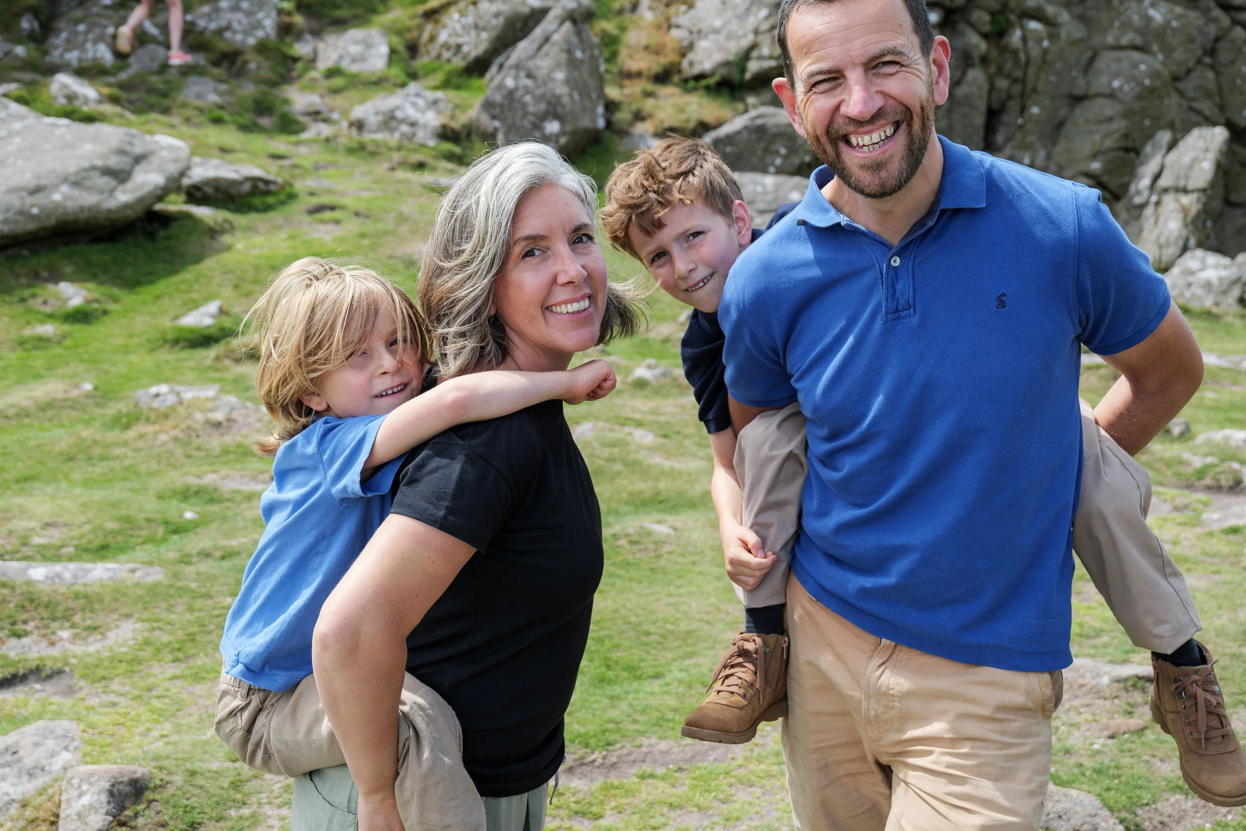 A smiling man in a blue shirt is carrying a young boy on his back, while a woman with gray hair and two children pose together outdoors near rocks and green grass.
