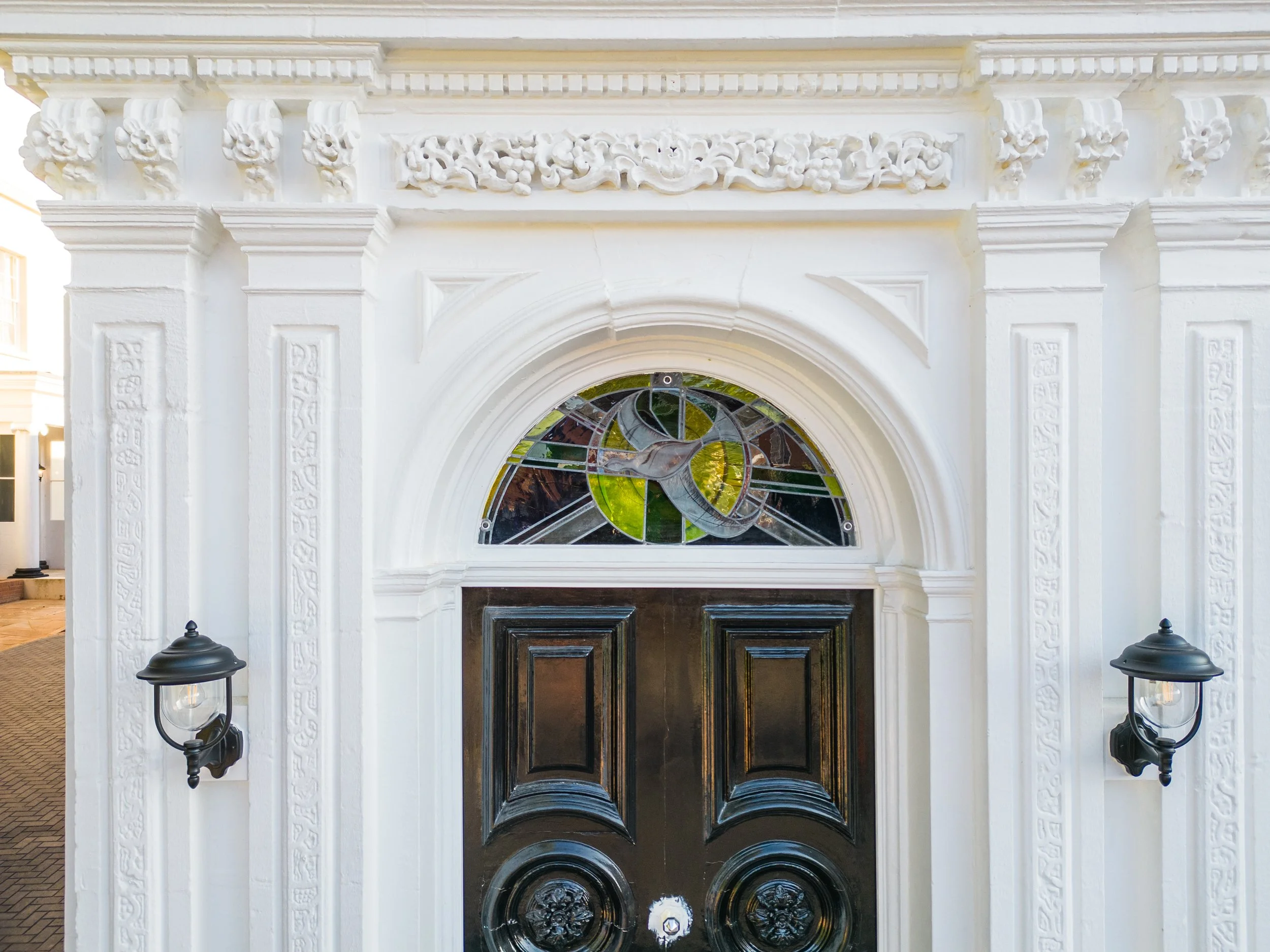 White decorative archway with intricate floral and scroll carvings, stained glass window with a spiral design, and two black outdoor lanterns on either side.