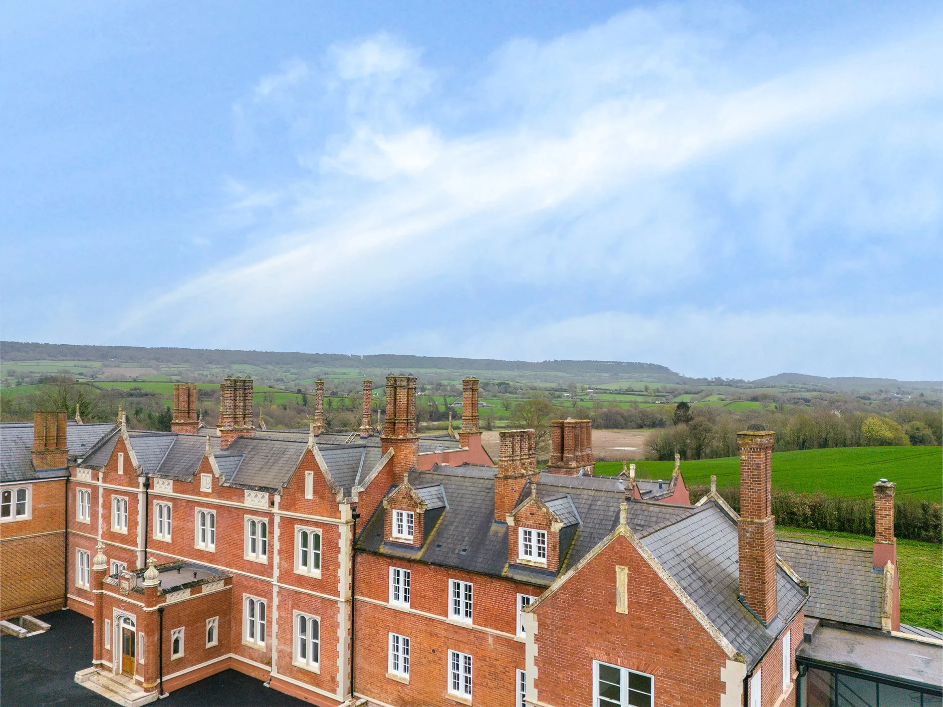 View of red brick residential buildings with chimneys, gray slate roofs, and white window frames, set against rolling green hills and a partly cloudy sky.