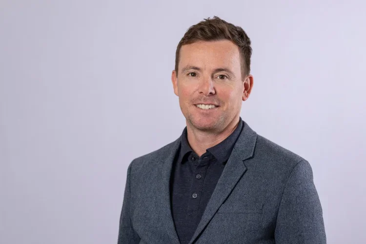 A man in a dark shirt and gray blazer smiling against a plain light background.