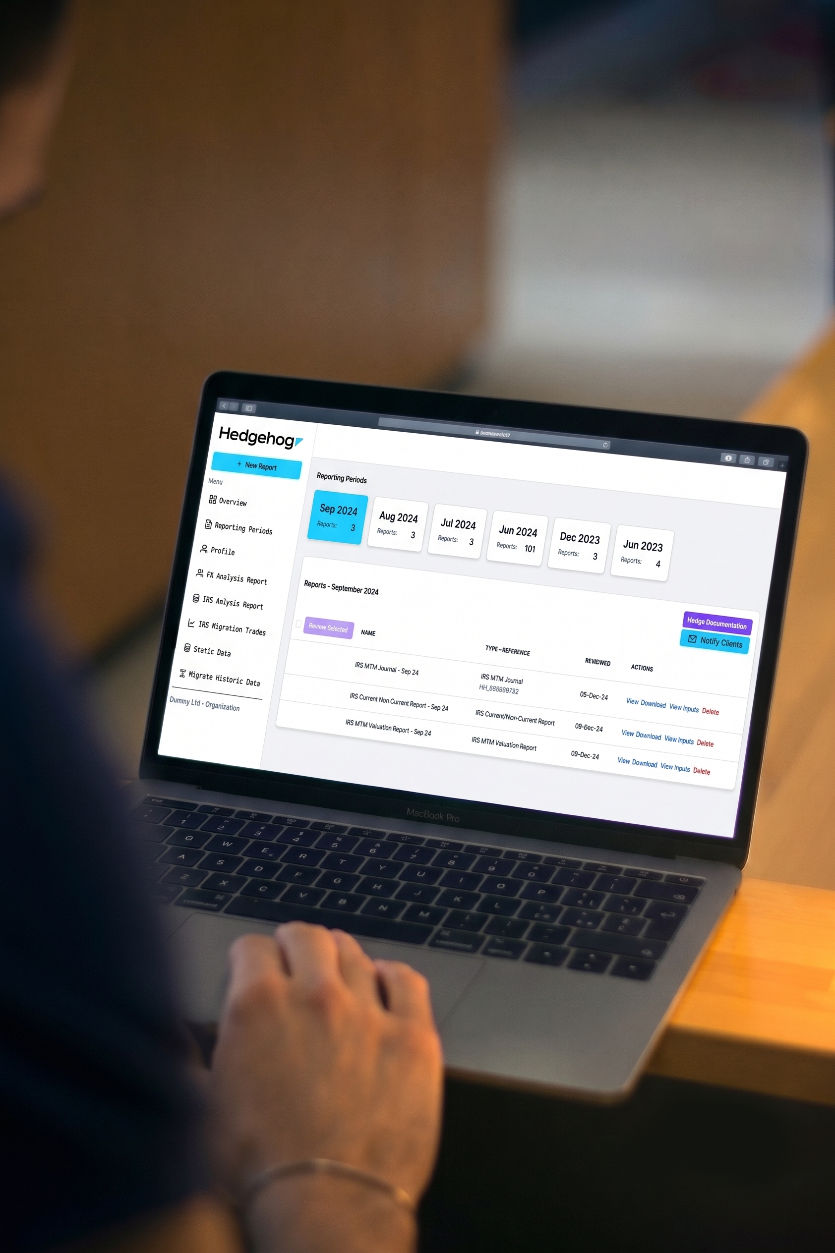 A person working on a MacBook Pro laptop displaying a report dashboard with menu options on the left, including Overview, Reporting Periods, Profile, and various analysis reports; person's hand visible near the keyboard, seated at a wooden table.