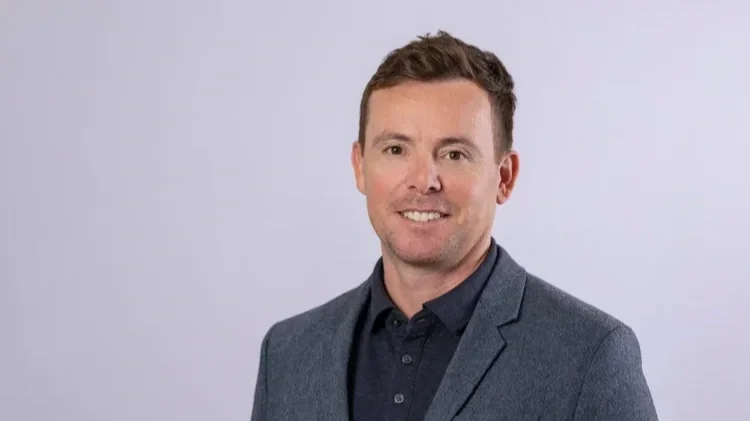 A smiling man with short brown hair, wearing a dark blue shirt and gray blazer, standing against a plain light gray background.