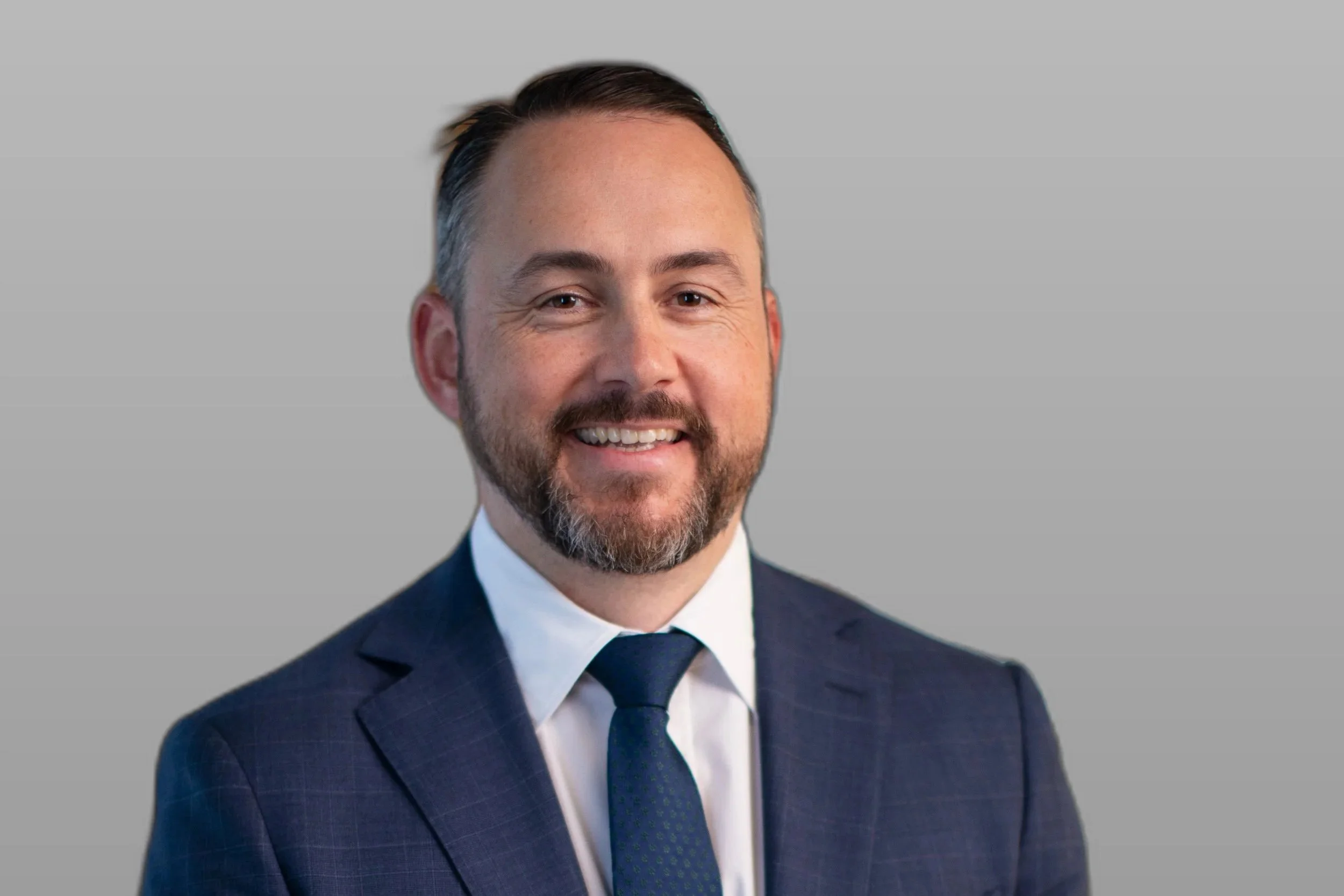 Headshot of a smiling man in a navy suit, white shirt, and blue tie, with short dark hair and a beard, against a gray background.