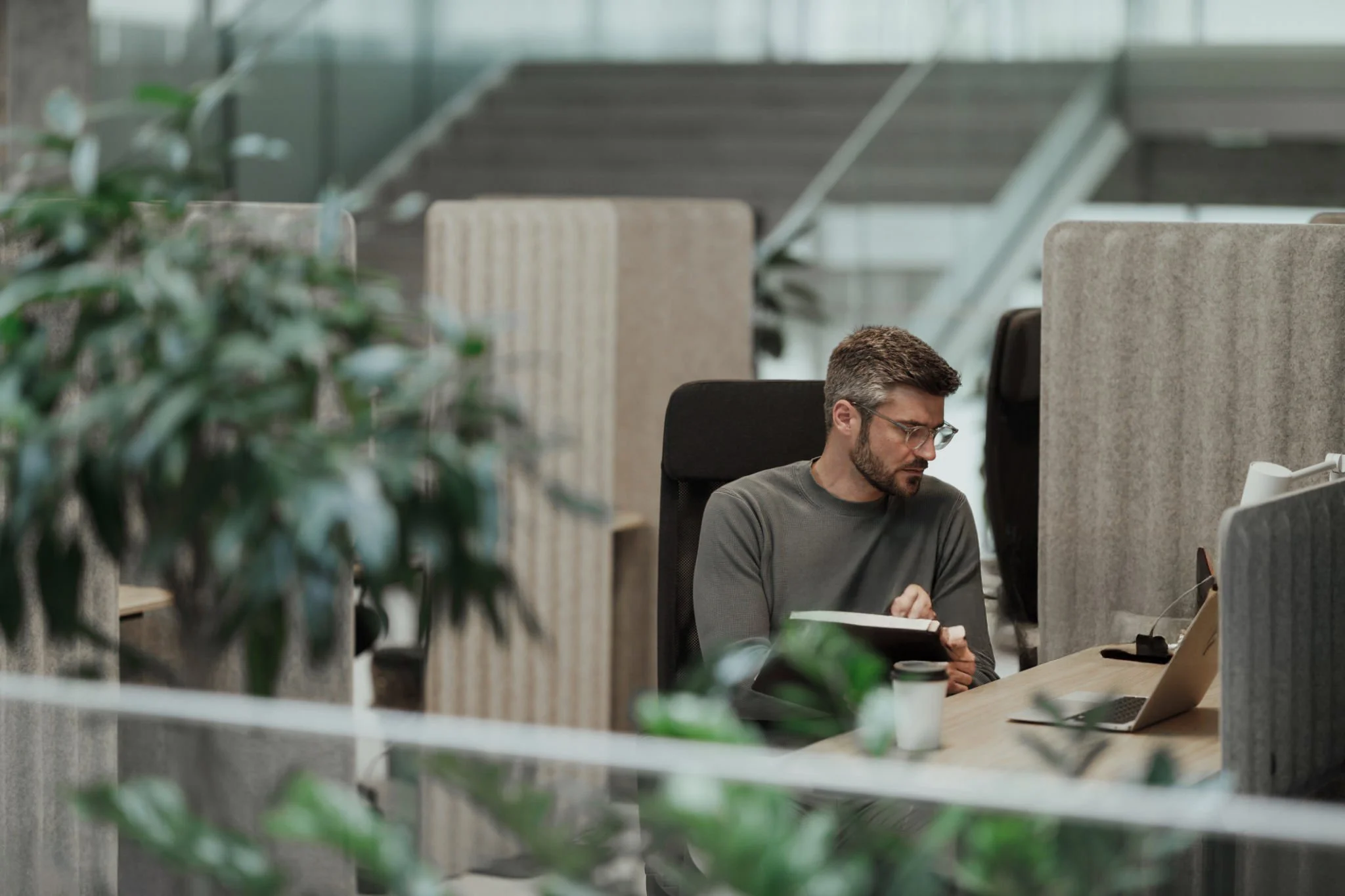 A man with glasses working on a computer in an office with glass walls, plants, and stairs in the background.