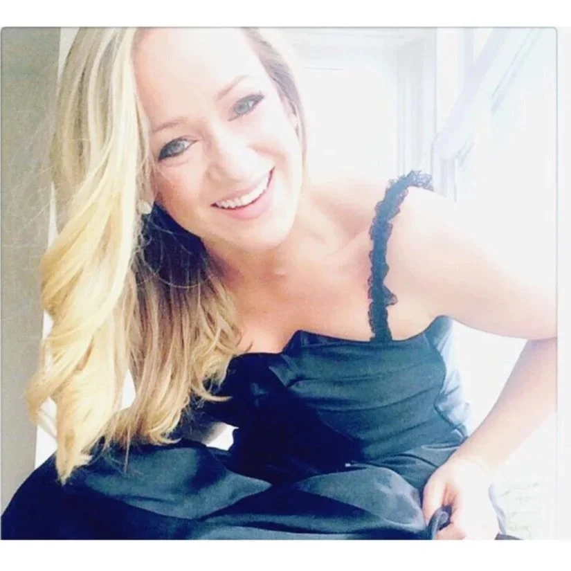 A smiling woman with long blonde hair, wearing a black dress with lace straps, leaning slightly forward and looking at the camera, with bright lighting from a window in the background.