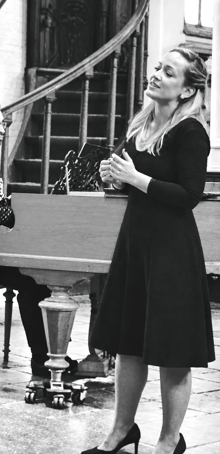 Black and white photo of a woman in a dark dress with high heels, standing beside a piano. She is smiling and holding a mug, appearing to be engaged in conversation. The background shows a staircase and part of a room with window and wooden details.