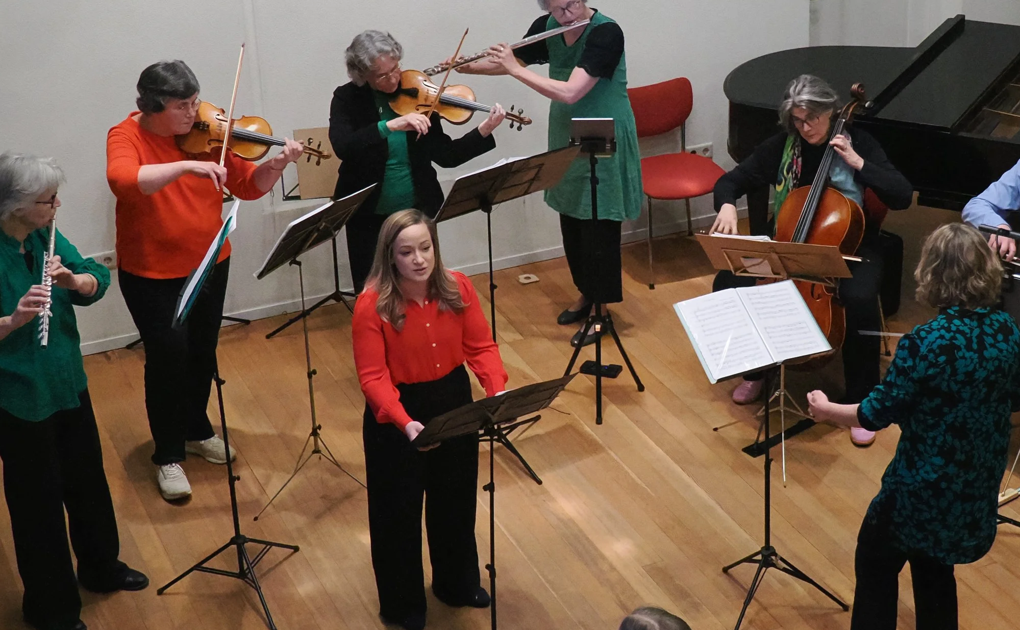 A group of musicians performing together indoors. There are people playing violins, a cello, a flute, and a piano, with sheet music on stands. Some members are standing, while others are sitting or leaning. The setting appears to be a small concert or rehearsal space with wooden flooring and simple decor.