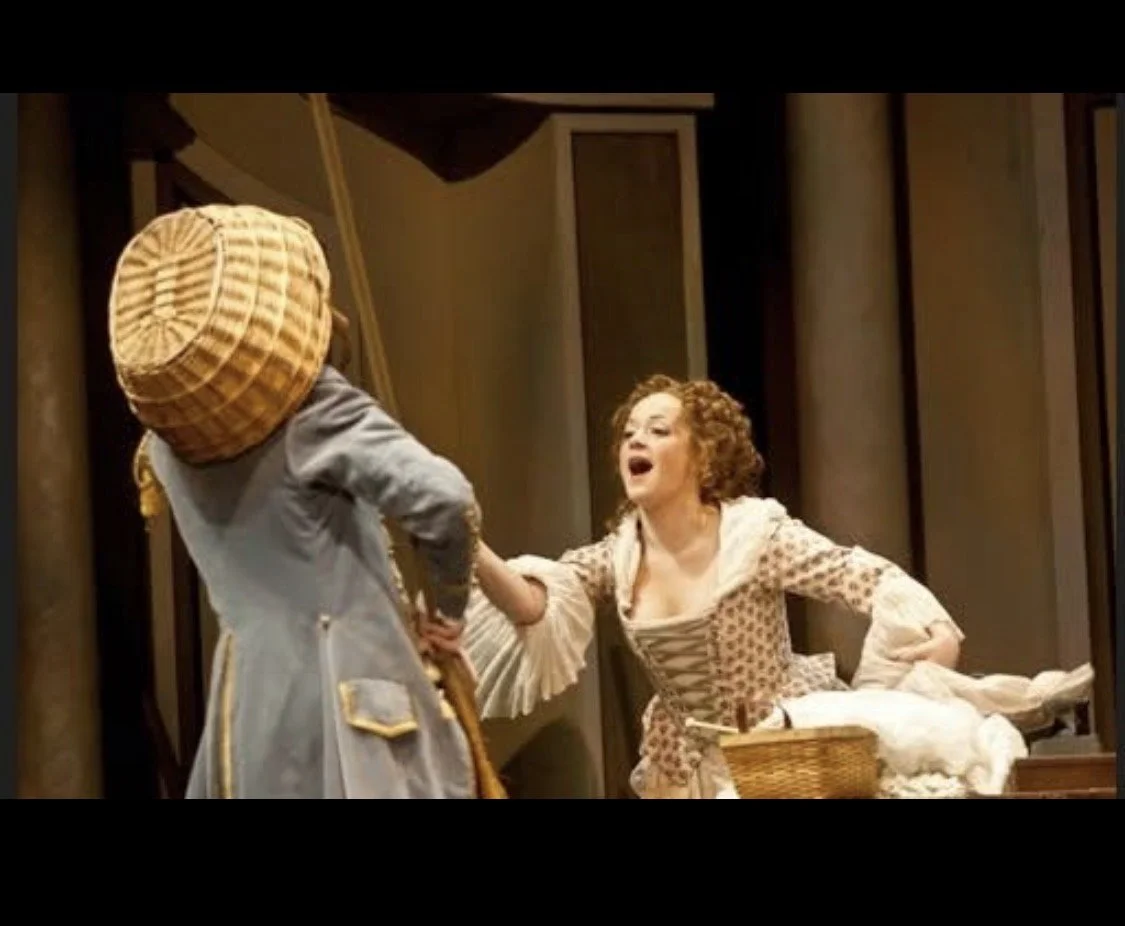 A theatrical scene with two women; one woman with curly hair is arguing with another woman wearing a bonnet, in a period costume, on a stage with wooden panels and a table.